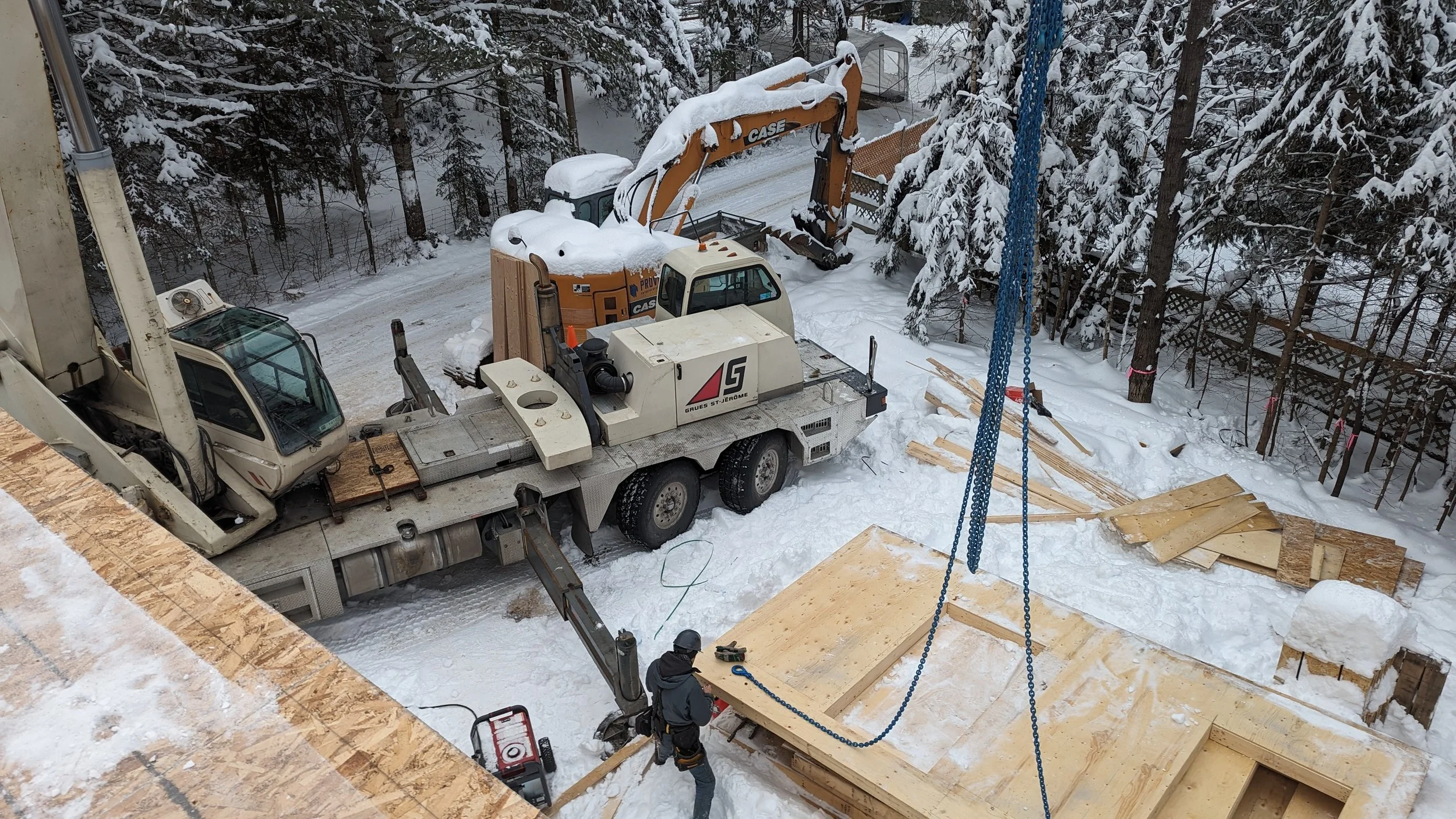 Construction site in snowy conditions with a crane lifting a wooden panel, a worker in a helmet and safety gear, a truck, a backhoe, and scattered wooden planks, surrounded by snow-covered trees.