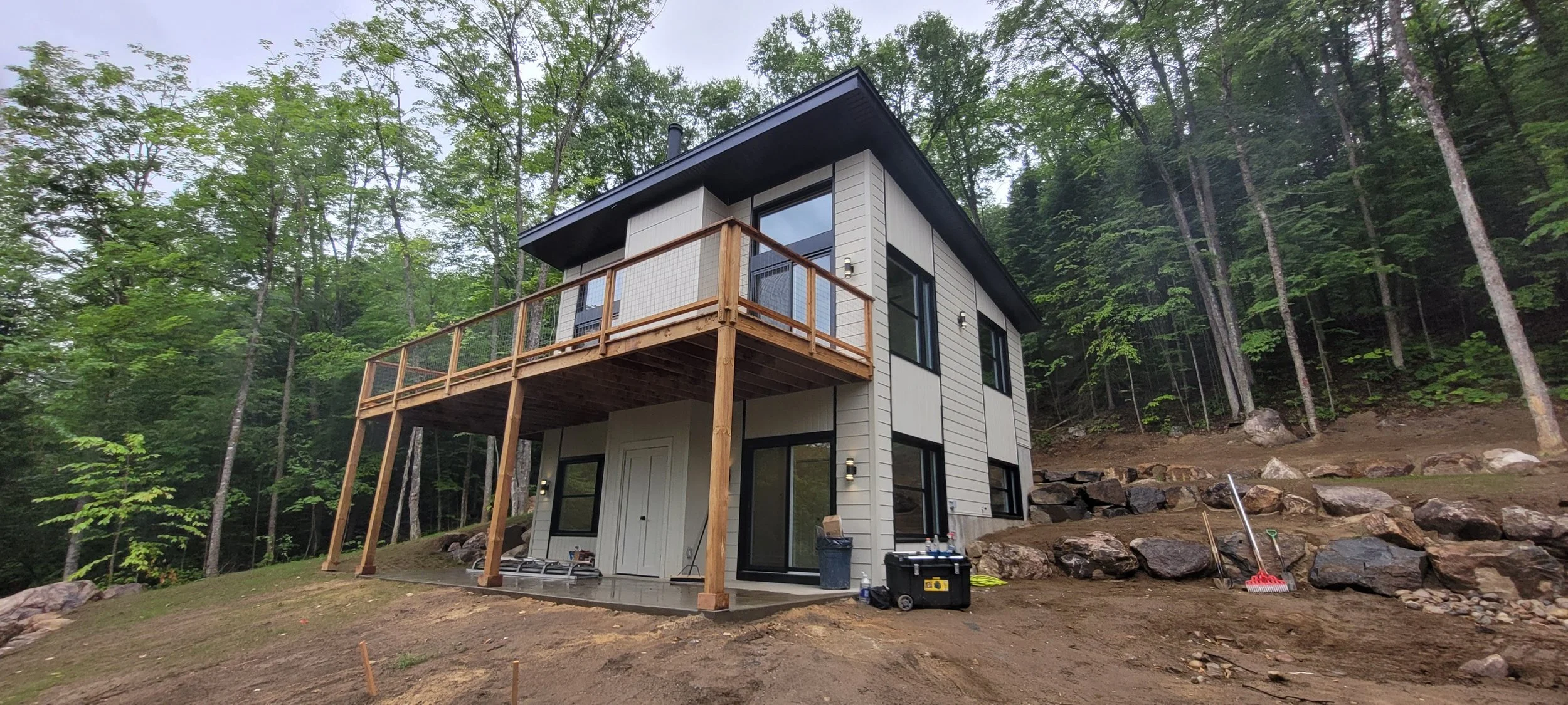 A modern three-story house with large windows and a balcony, surrounded by trees and rocks, with construction tools and equipment on the ground.