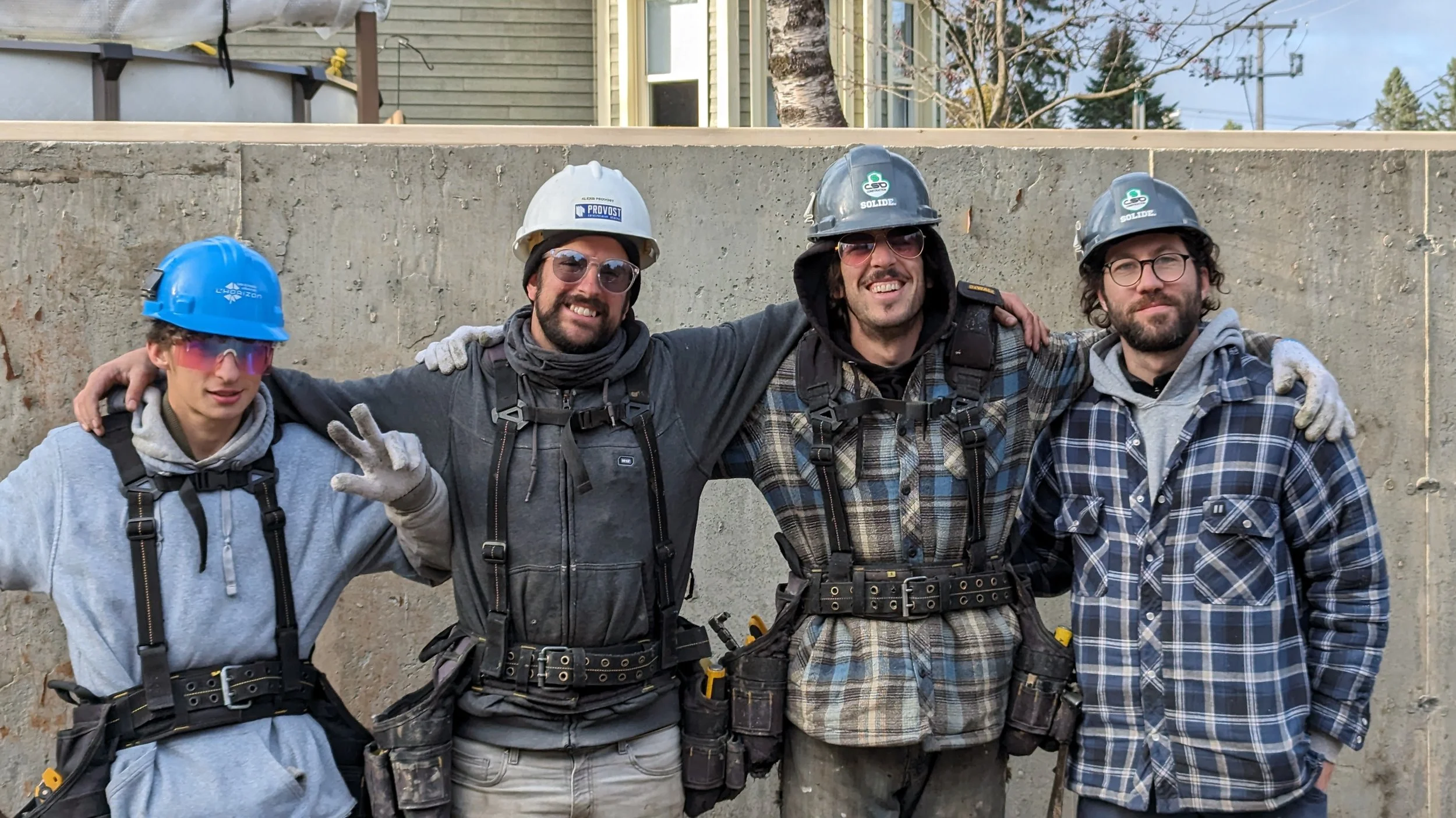 Four construction workers standing side by side with arm around each other in front of a concrete wall. They are all wearing helmets, safety gloves, and casual work clothes. The workers are smiling and looking at the camera.