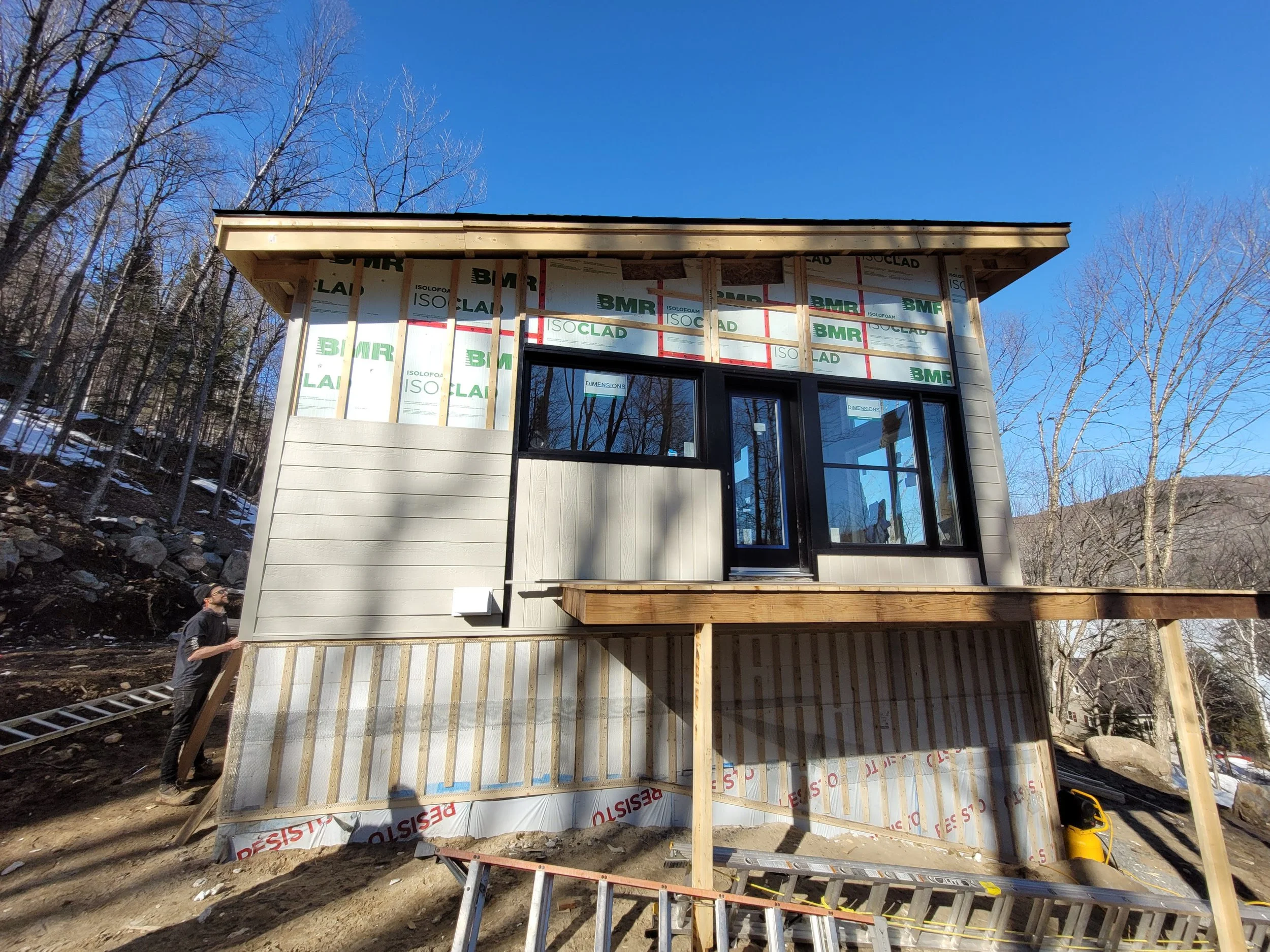 Construction of a house on a hill with a partially finished exterior, large windows, and a wooden deck, surrounded by leafless trees under a clear blue sky.