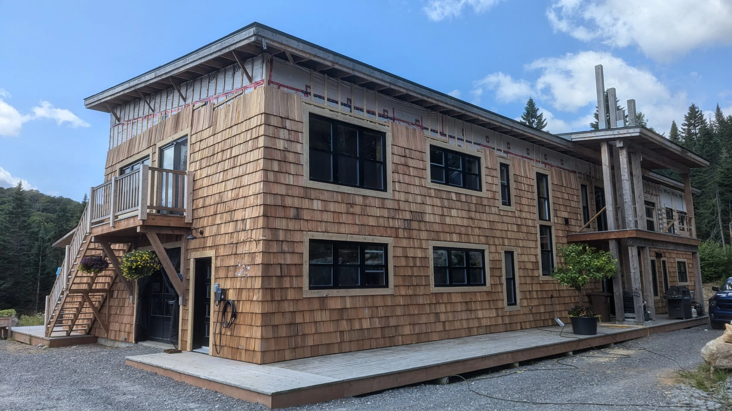 Under construction wooden multi-unit residential building with exterior shingle siding, multiple windows, small balcony, and outdoor staircase, set in a rural area with trees in the background and a gravel pathway in front.