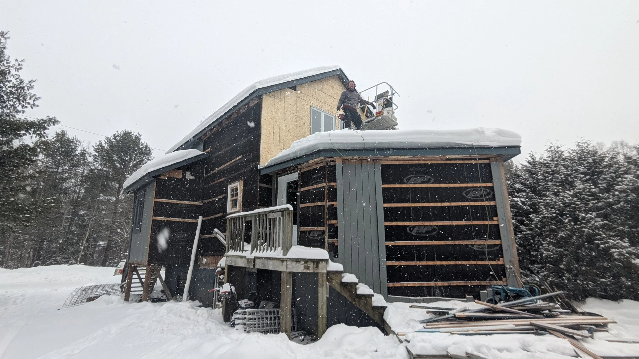 A house under construction during snowfall, with a person standing on a lift on the roof, working on the exterior wall.