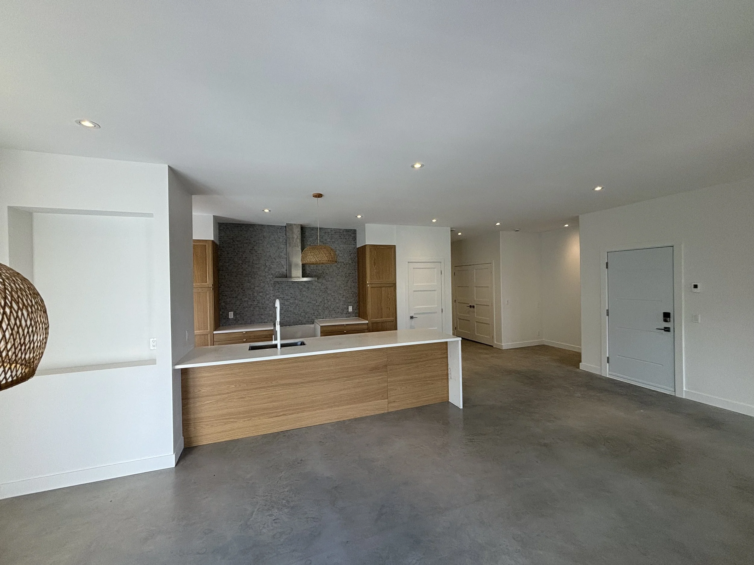 Empty modern kitchen with white walls, wooden cabinets, and a kitchen island with a white countertop, gray tiled backsplash, and a stainless steel range hood. Open living space with concrete floors and multiple ceiling lights.
