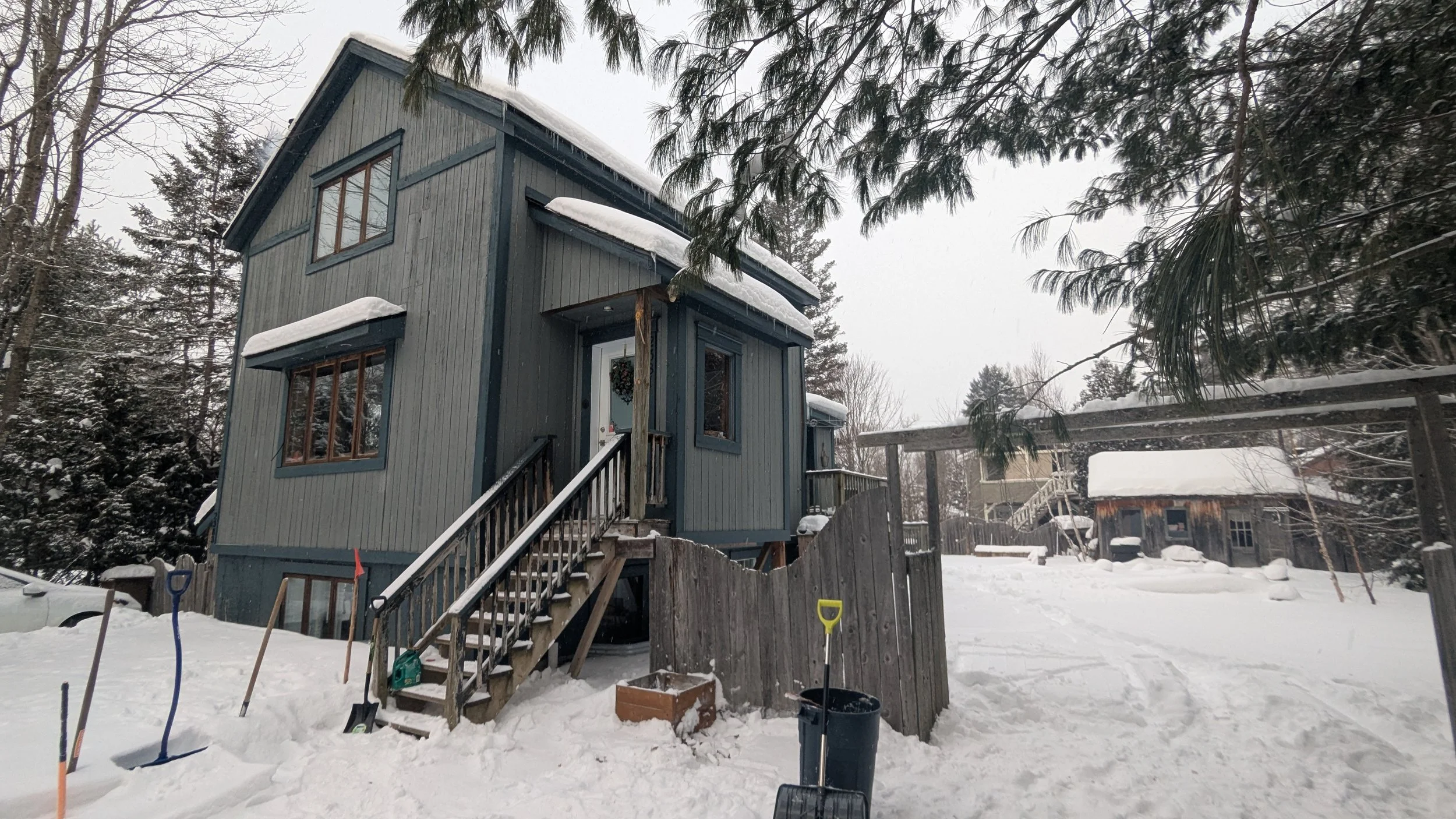 A blue and gray two-story wooden house with a snow-covered roof is surrounded by snow and trees, with a wooden staircase leading to the front door, and snow shovels and a black trash can in the yard.
