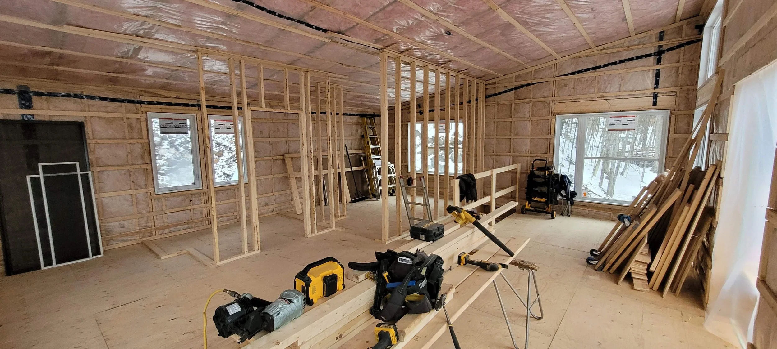 Interior of a house under construction with exposed wooden framing, insulation on the ceiling, windows, and various construction tools and materials scattered around.