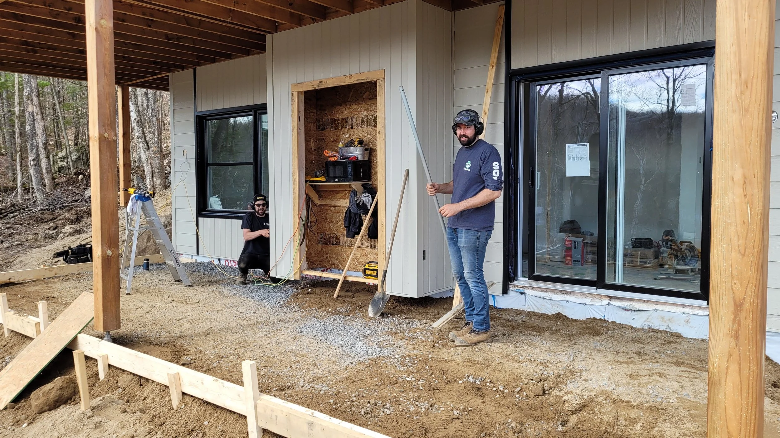 Two men are working on the construction of a house's exterior wall. One man is standing with a measuring pole, while the other is squatting near an open wall space with tools and materials around him. The house has sliding glass doors and is surrounded by a wooded area.