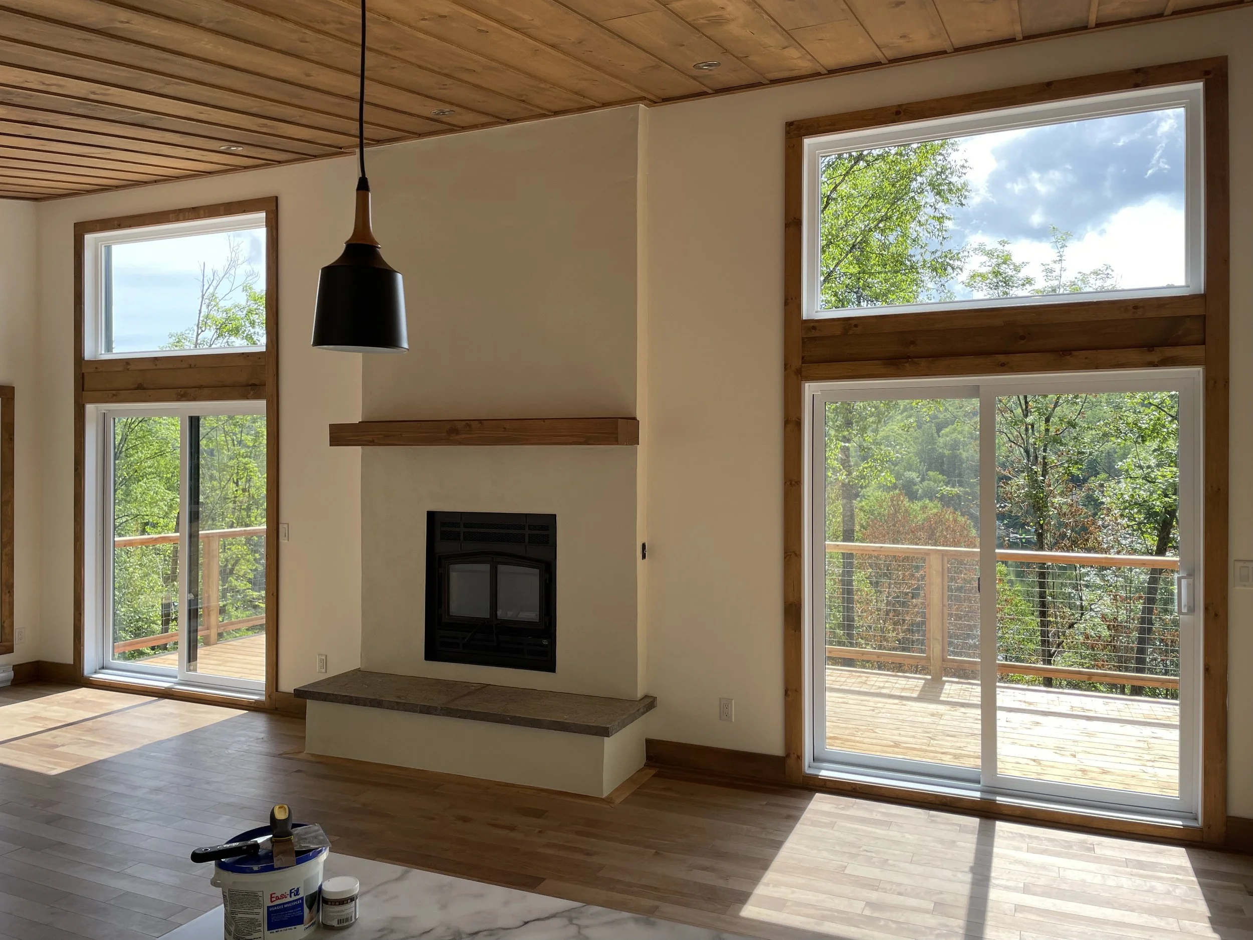 Interior view of a living room with large windows and sliding glass door leading to a deck, wooden ceiling, fireplace, and hardwood floors with paint cans and tools on a nearby surface.