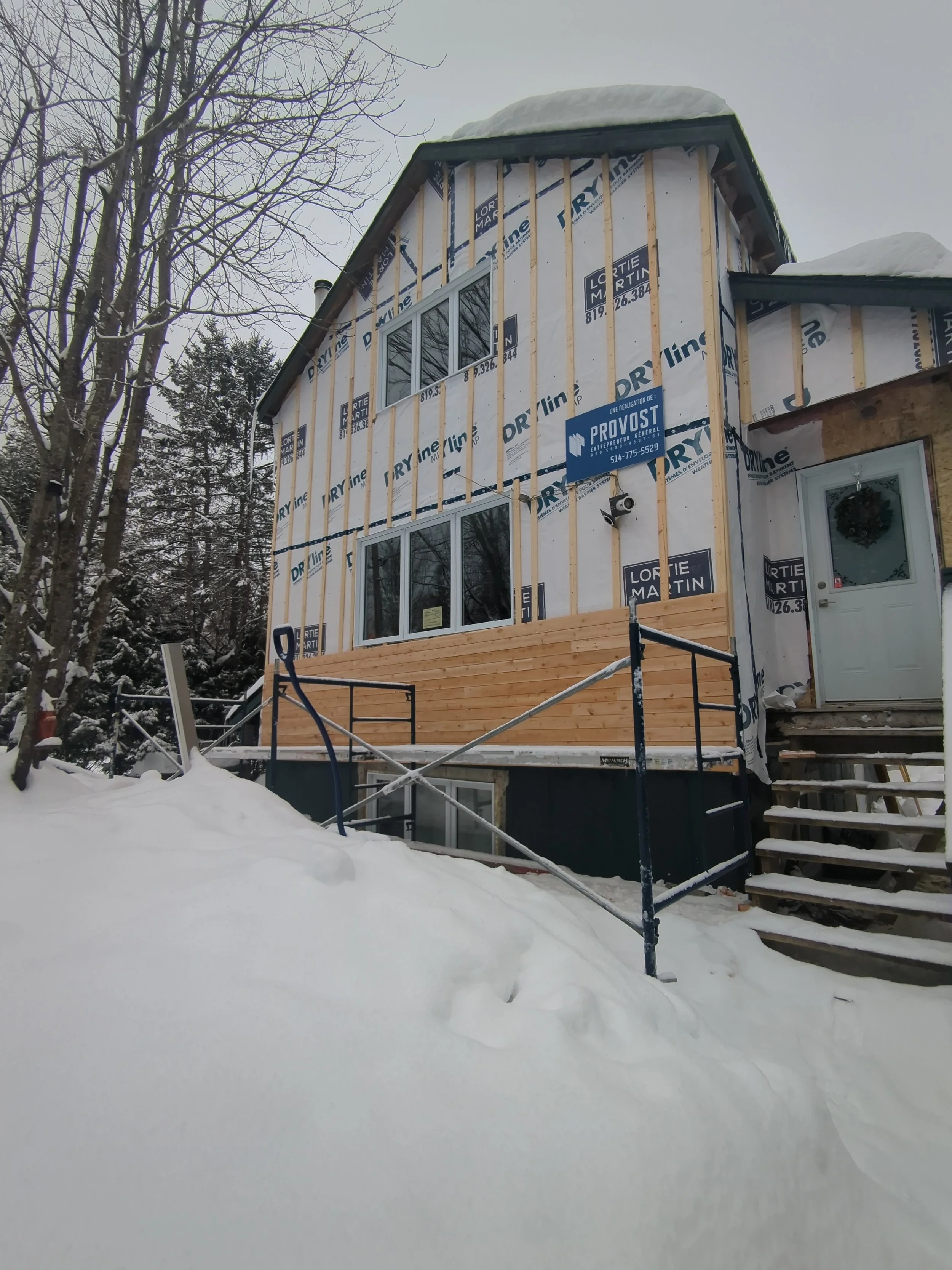 A house under construction with snow on the ground and on the roof, with visible wood siding and windows, surrounded by trees in winter.