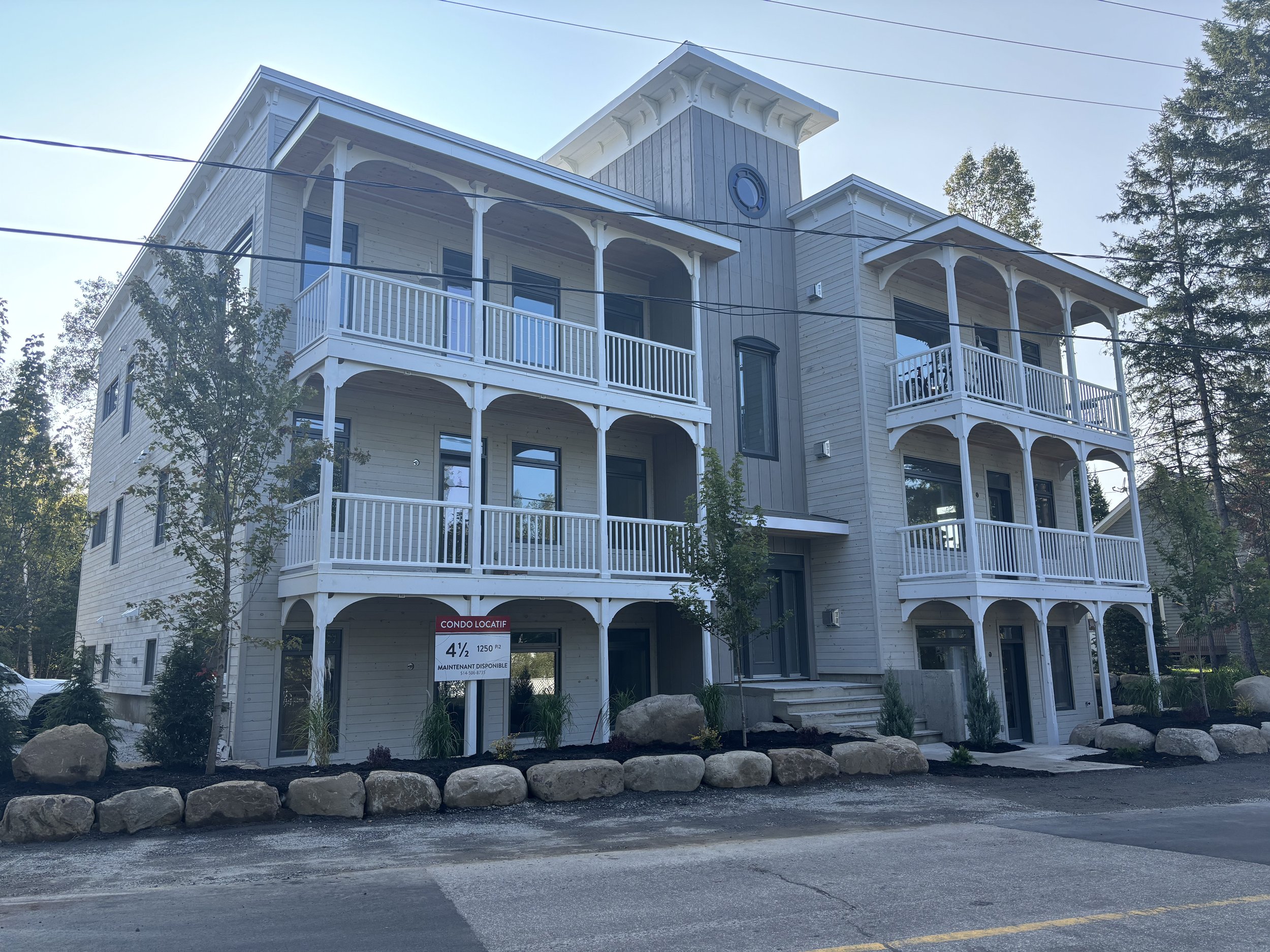 Multi-story residential building with balconies and a sign in front indicating availability for sale or rent, surrounded by trees and large rocks.