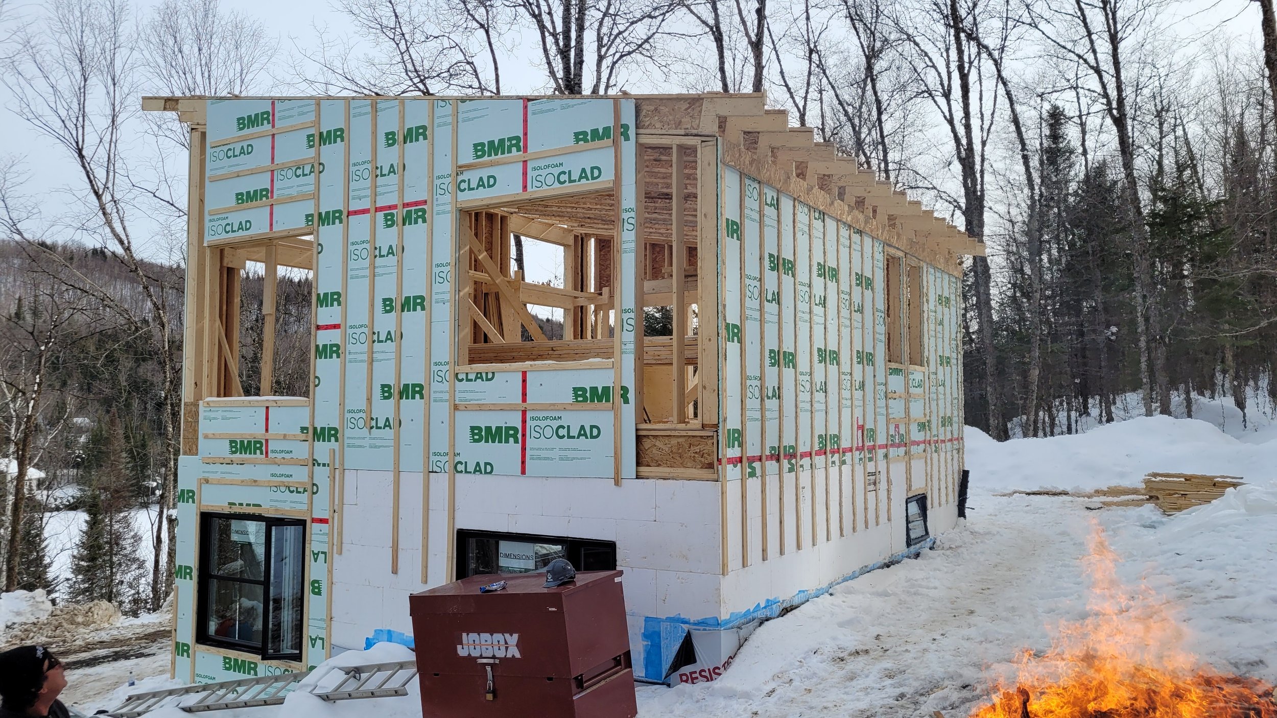 A house under construction surrounded by snow, with a fire burning in the foreground, and trees in the background.