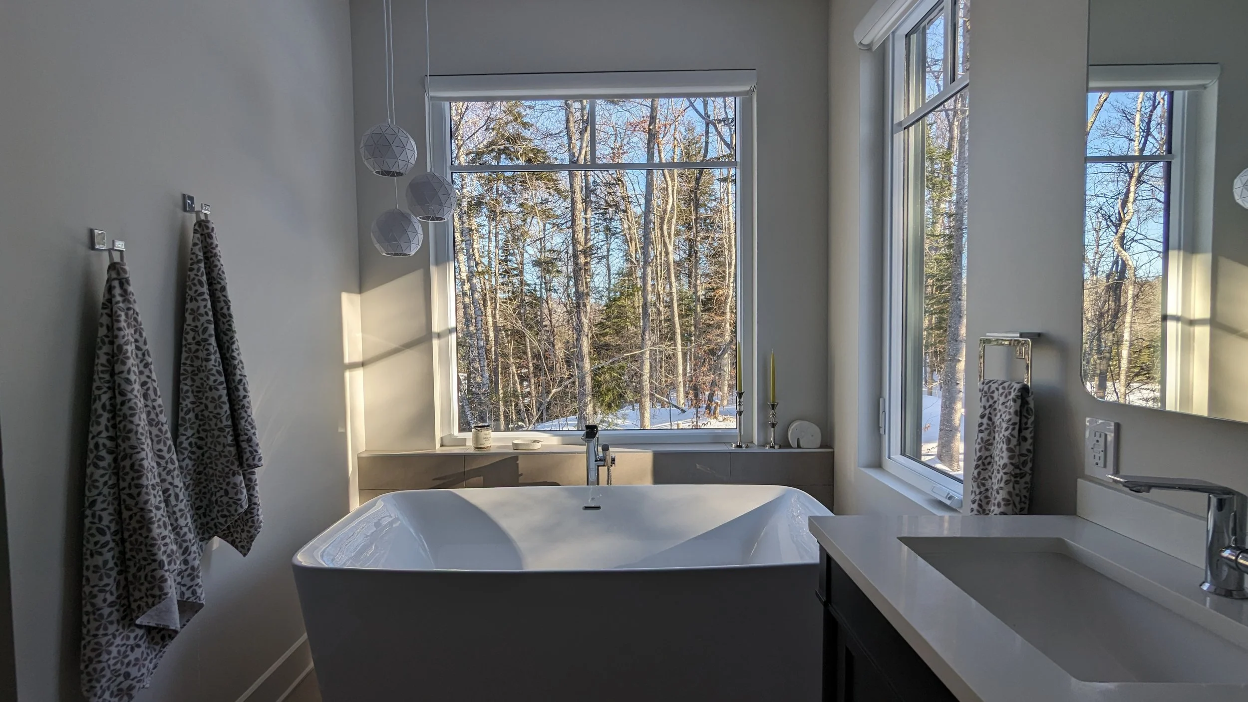 Modern bathroom with large window overlooking a snowy wooded landscape, featuring a freestanding bathtub, twin candles, and towels hanging on hooks