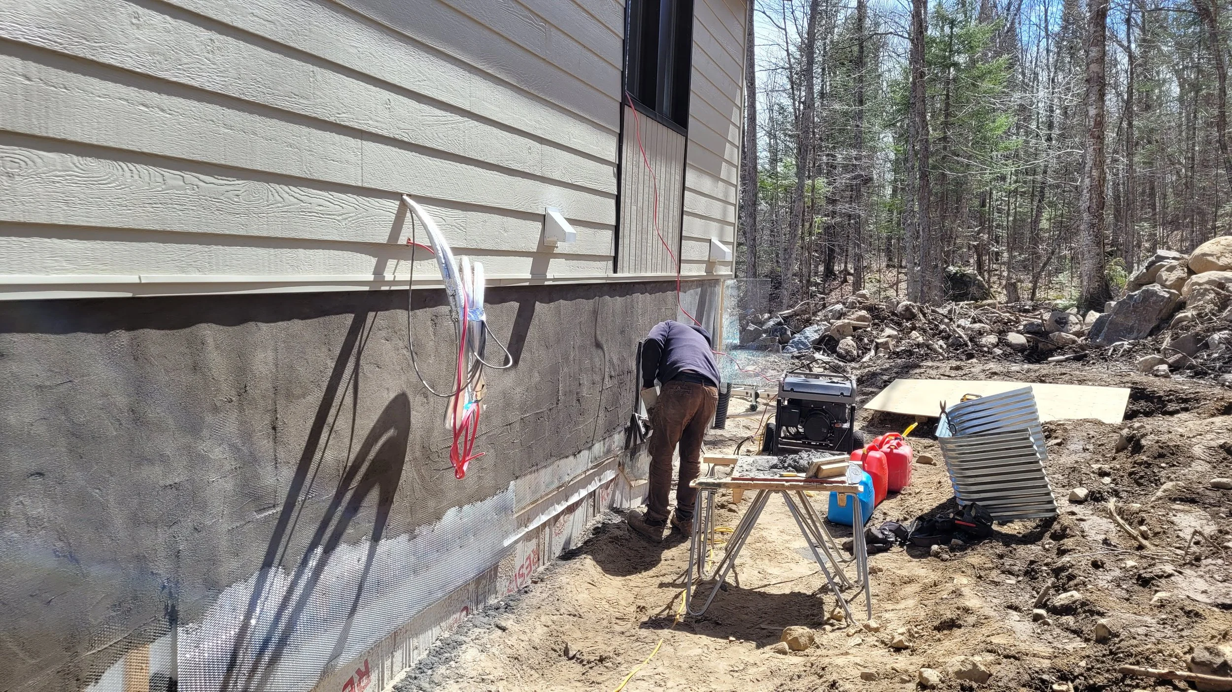 Construction worker installing insulation on the foundation of a house, with building materials and equipment around, in a wooded outdoor area.