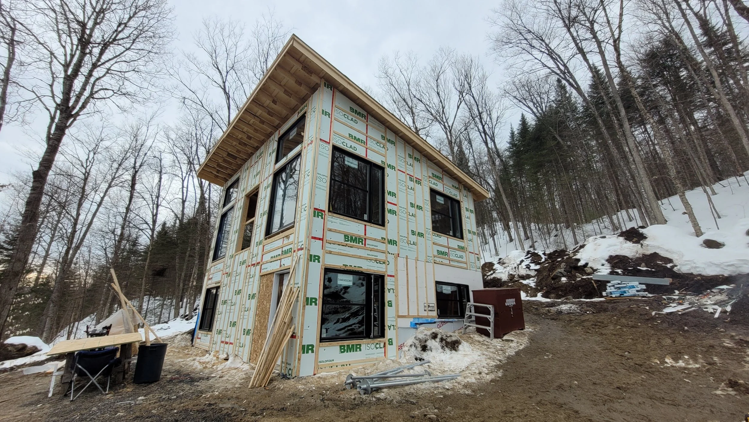 A two-story house under construction on a snowy hillside, with the exterior wall insulation and framing visible, surrounded by trees.