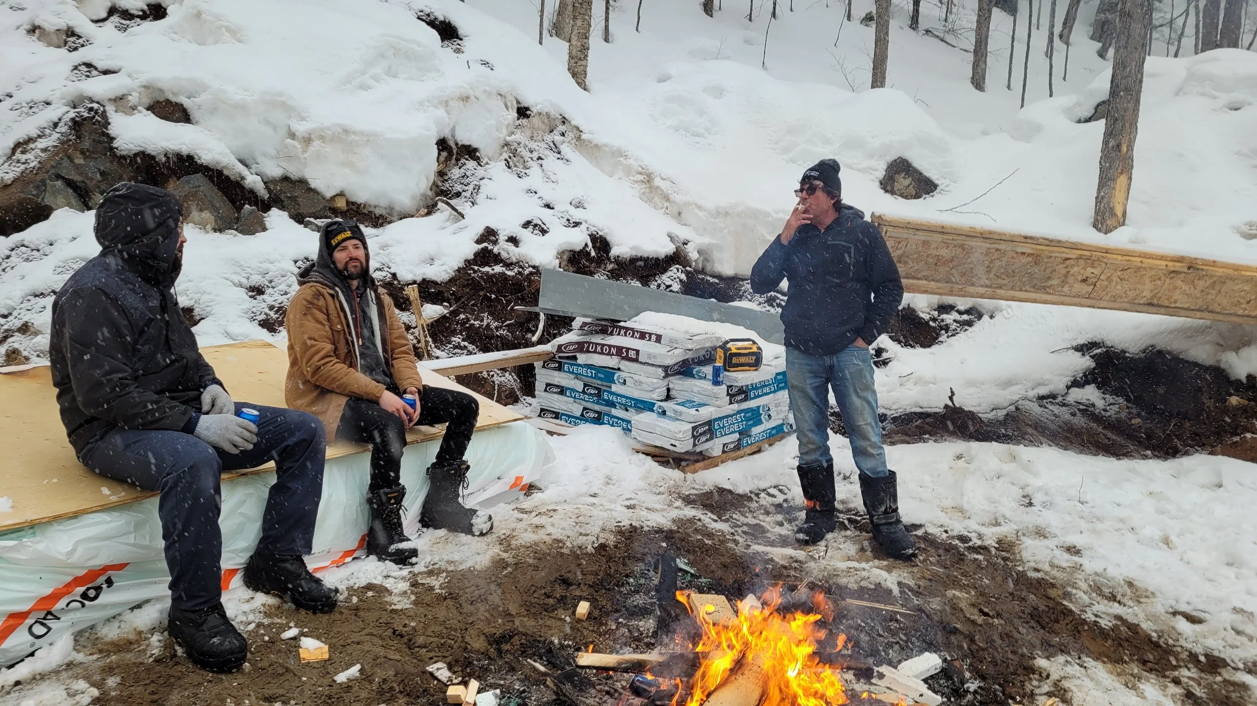 Three men in winter clothing sitting and standing around a small campfire in a snowy outdoor setting with construction materials nearby.