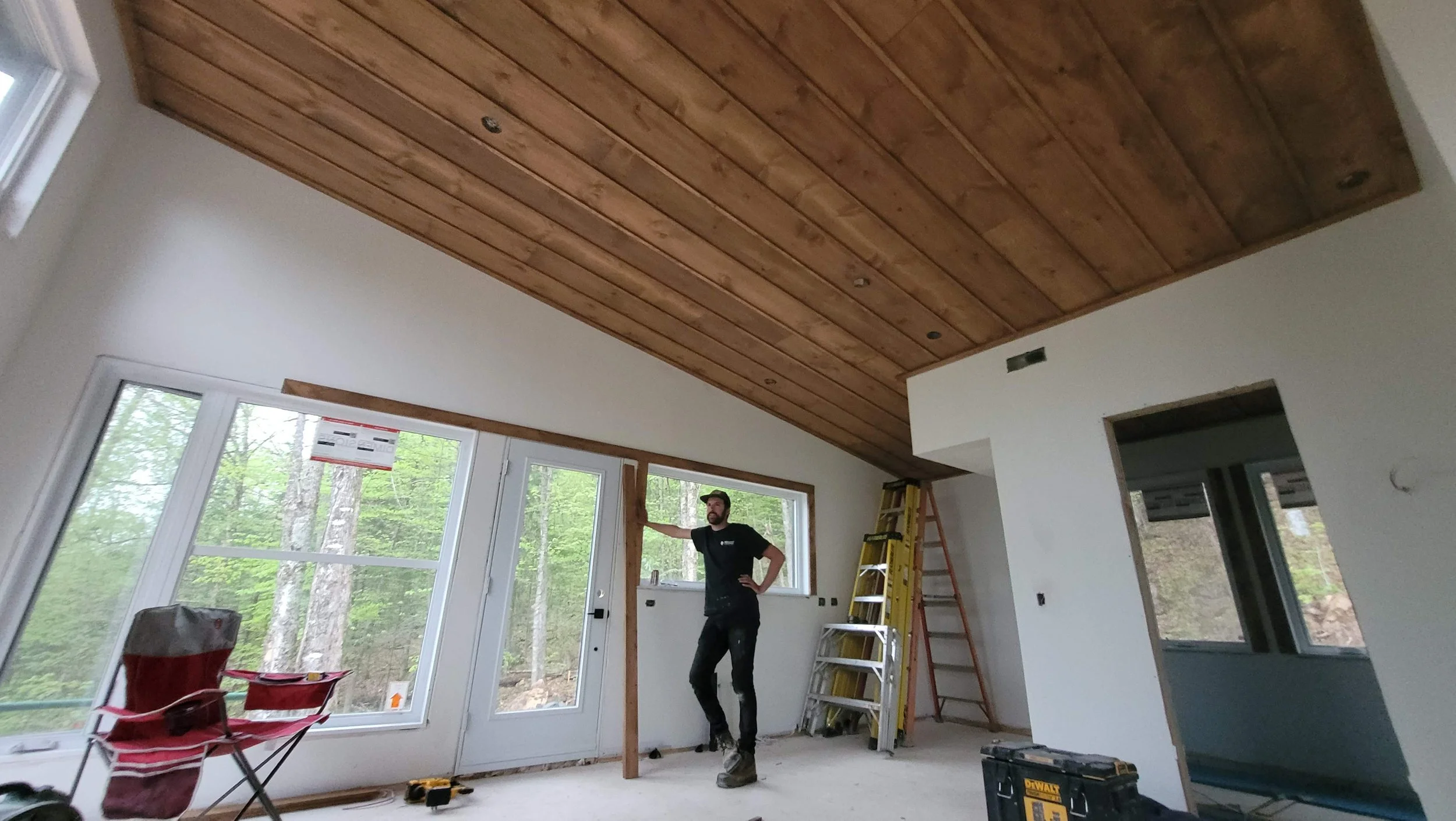 Interior of a room under construction with a man standing near a window and a wooden beam on the wall, with ladders and construction tools present.