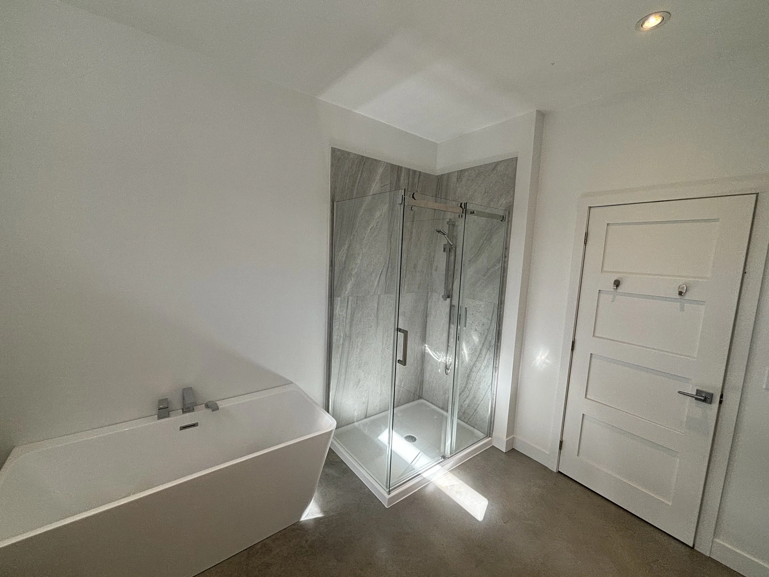 A modern bathroom with a white bathtub, a glass-enclosed shower with gray marble tiles, and a white door.