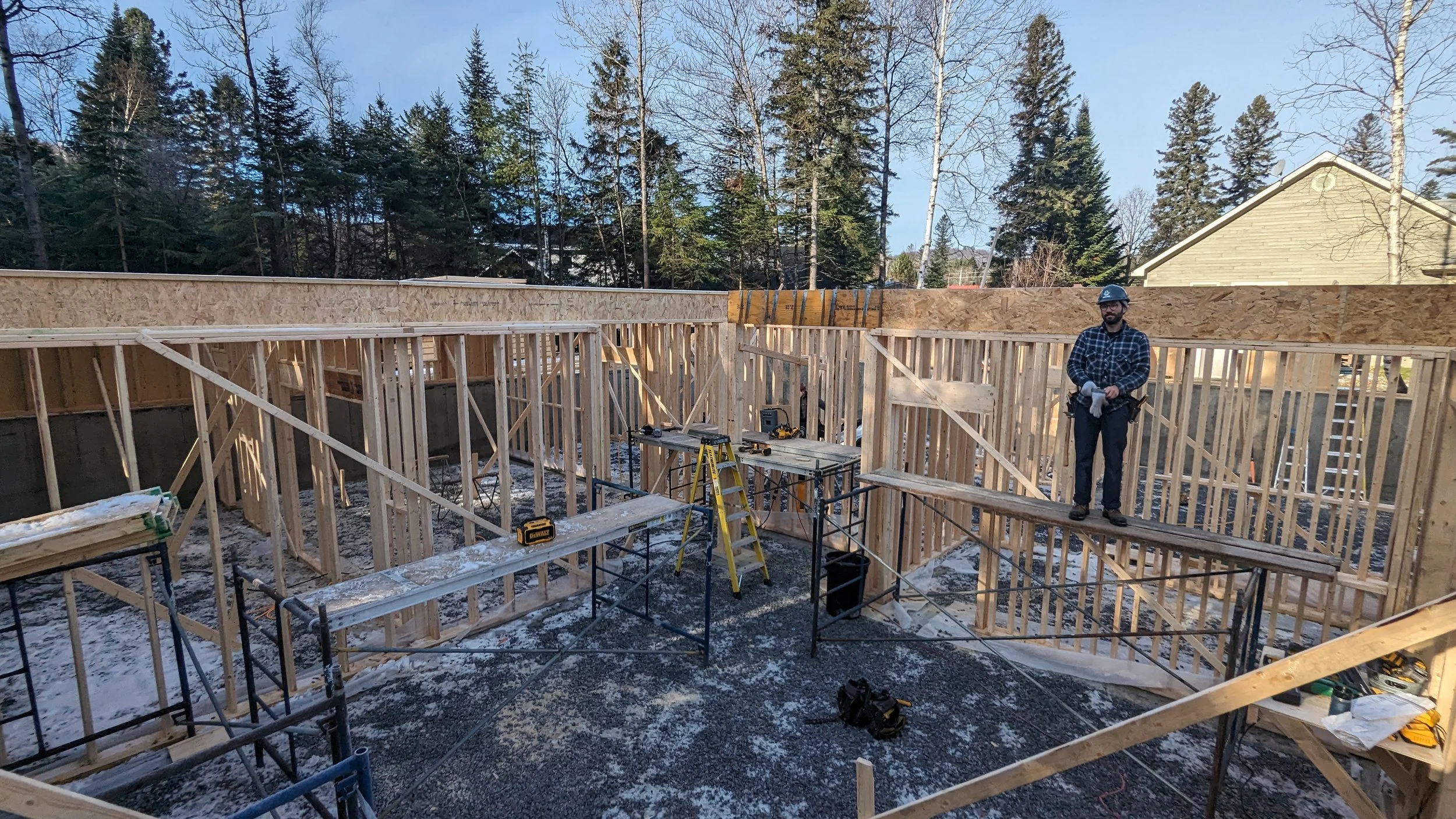 Construction site with wooden framing for a building, a worker standing on a plank, tools on tables, surrounded by scaffolding, outdoors with trees and a house in the background.