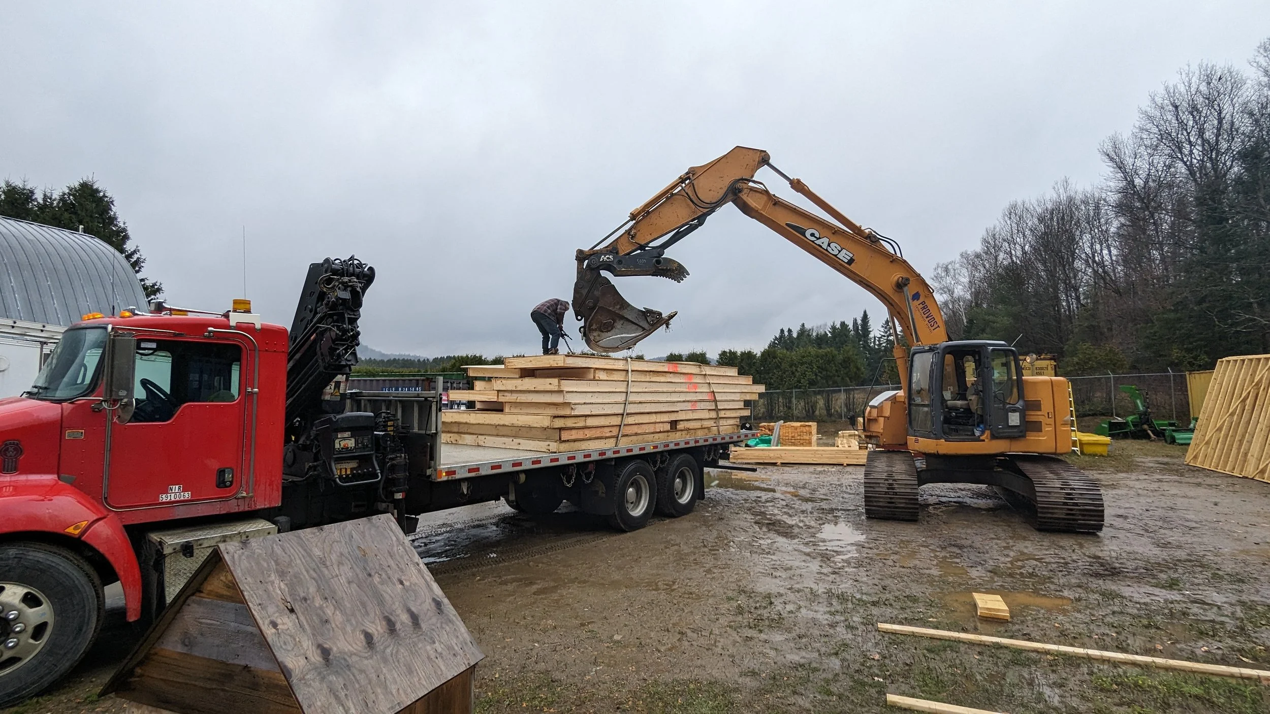 Construction site with a red truck and an orange excavator loading wooden planks, overcast sky, muddy ground, trees in the background.