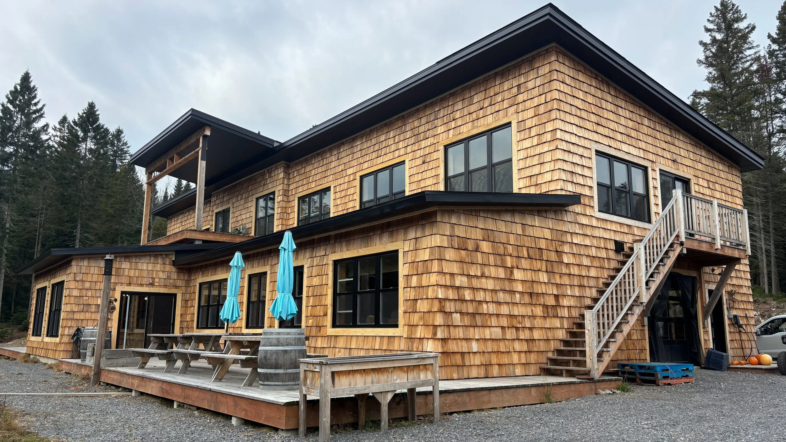 A two-story house with a wooden shingle exterior, black framed windows, a small balcony, and an outdoor deck with picnic tables and umbrellas, situated in a wooded area.