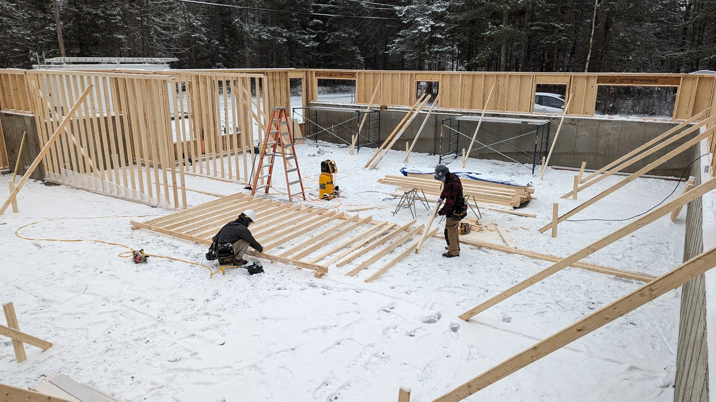 Two construction workers building a wooden frame house in snowy weather, surrounded by snow and construction tools.