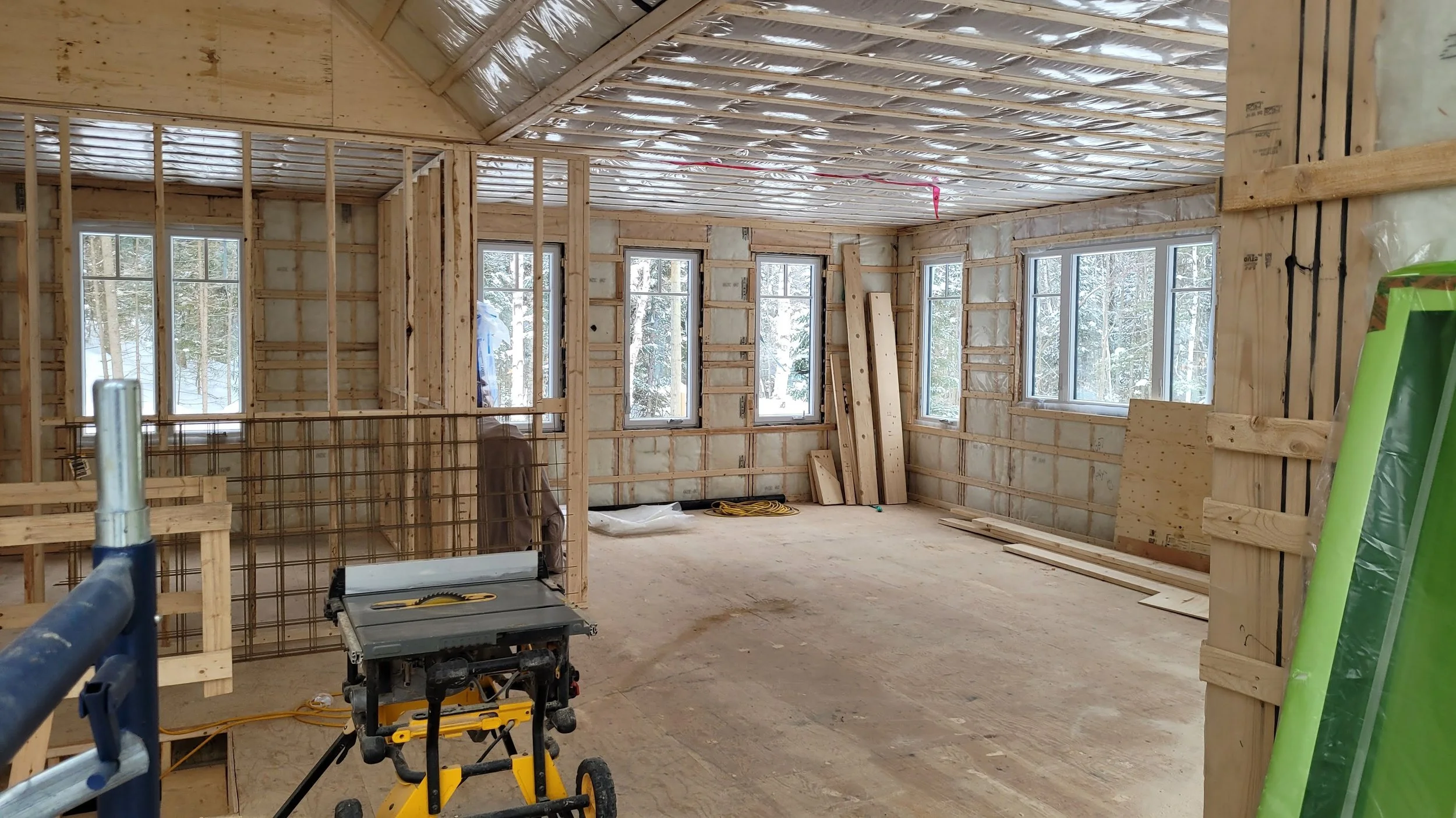 Interior of a house under construction with wooden framing, multiple windows, and construction tools on the floor.