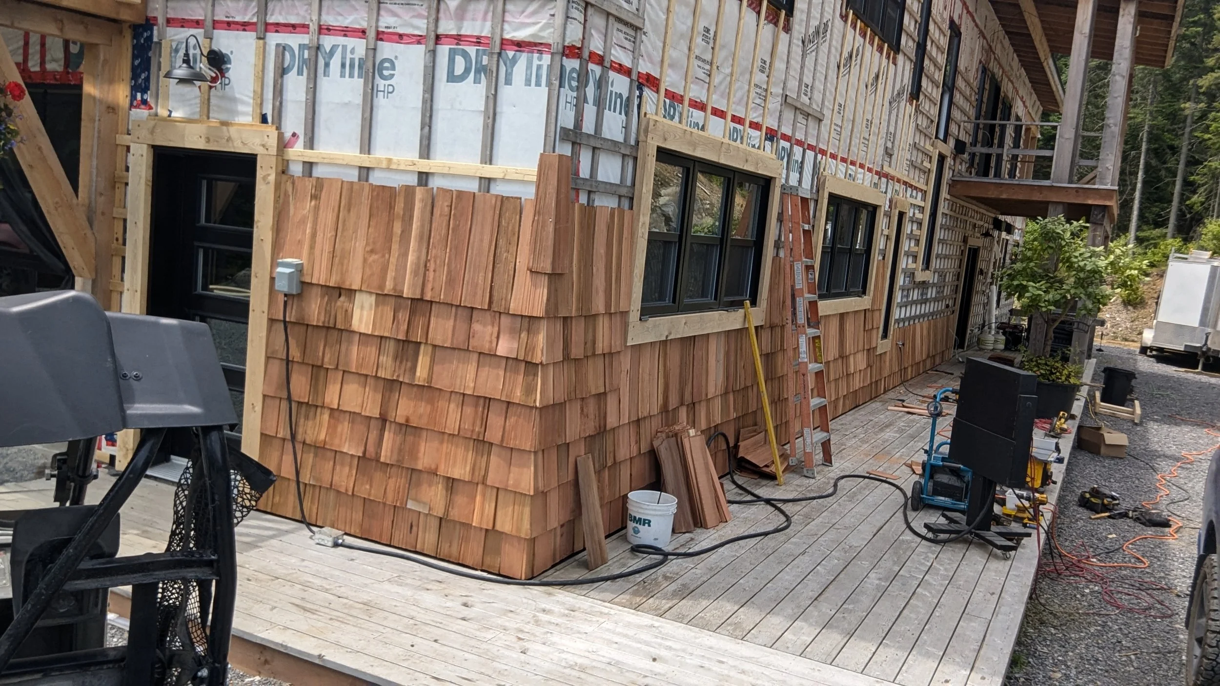 Construction of a house with wooden siding, black window frames, and a wooden deck in progress, with construction tools and materials around.