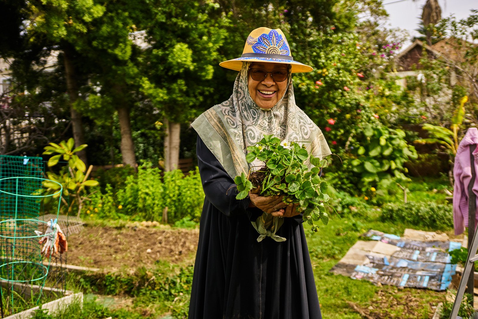 Photo of person in hijab and sun hat holding plants and smiling in a community garden
