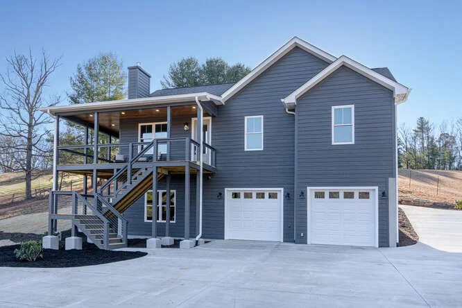 A gray two-story house with a double garage, a porch with stairs, and a balcony, set in a suburban area with trees.