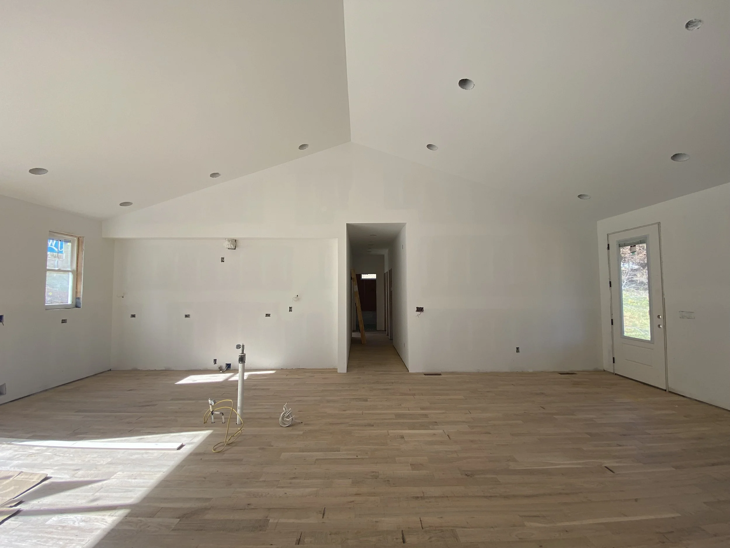 Empty room undergoing construction with white walls, wooden flooring, small windows, and a door, with electrical work in progress.