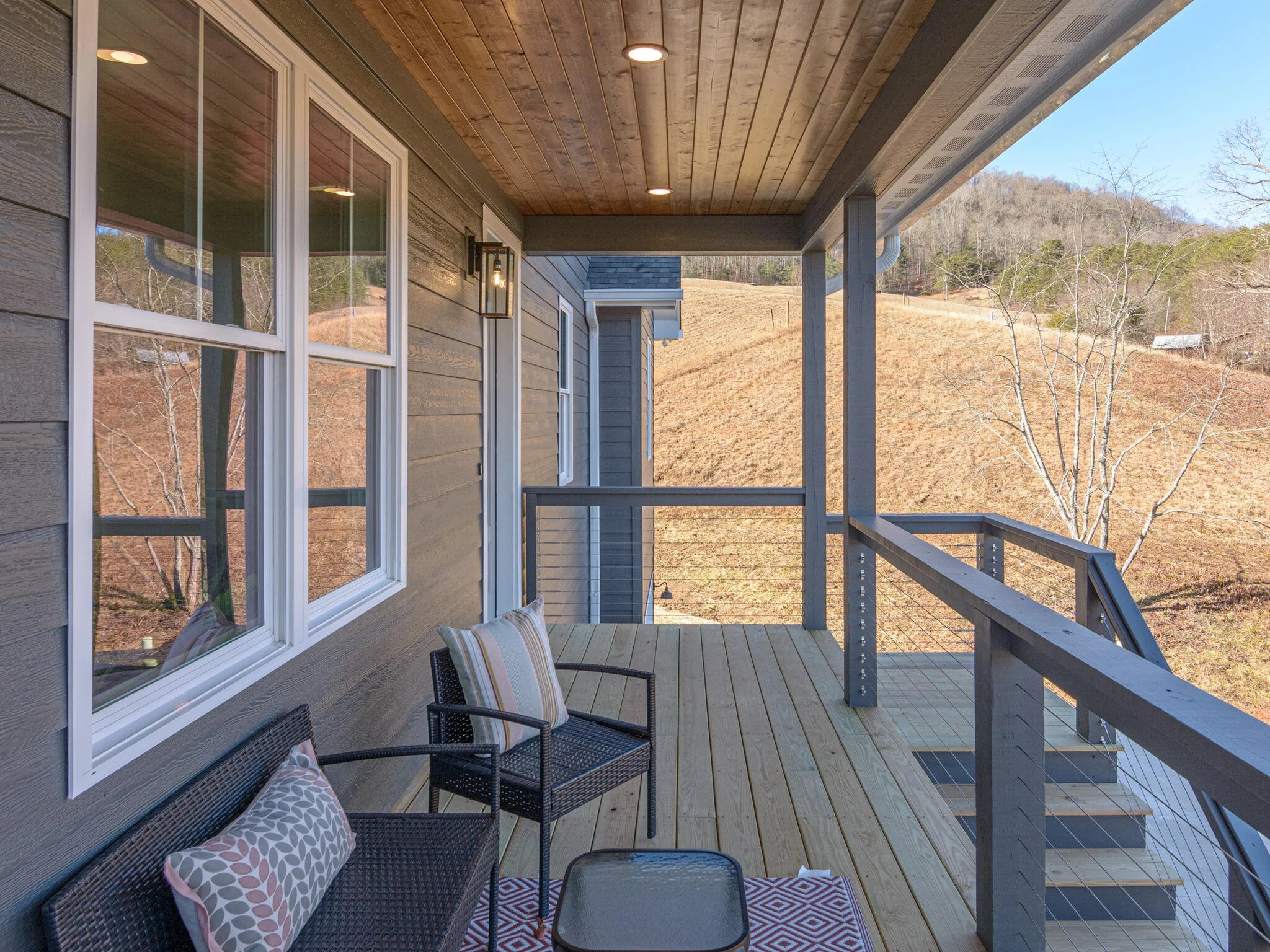 View from a house balcony overlooking a hillside with sparse trees and grass, with outdoor seating including two chairs and a small table, and some decorative pillows.