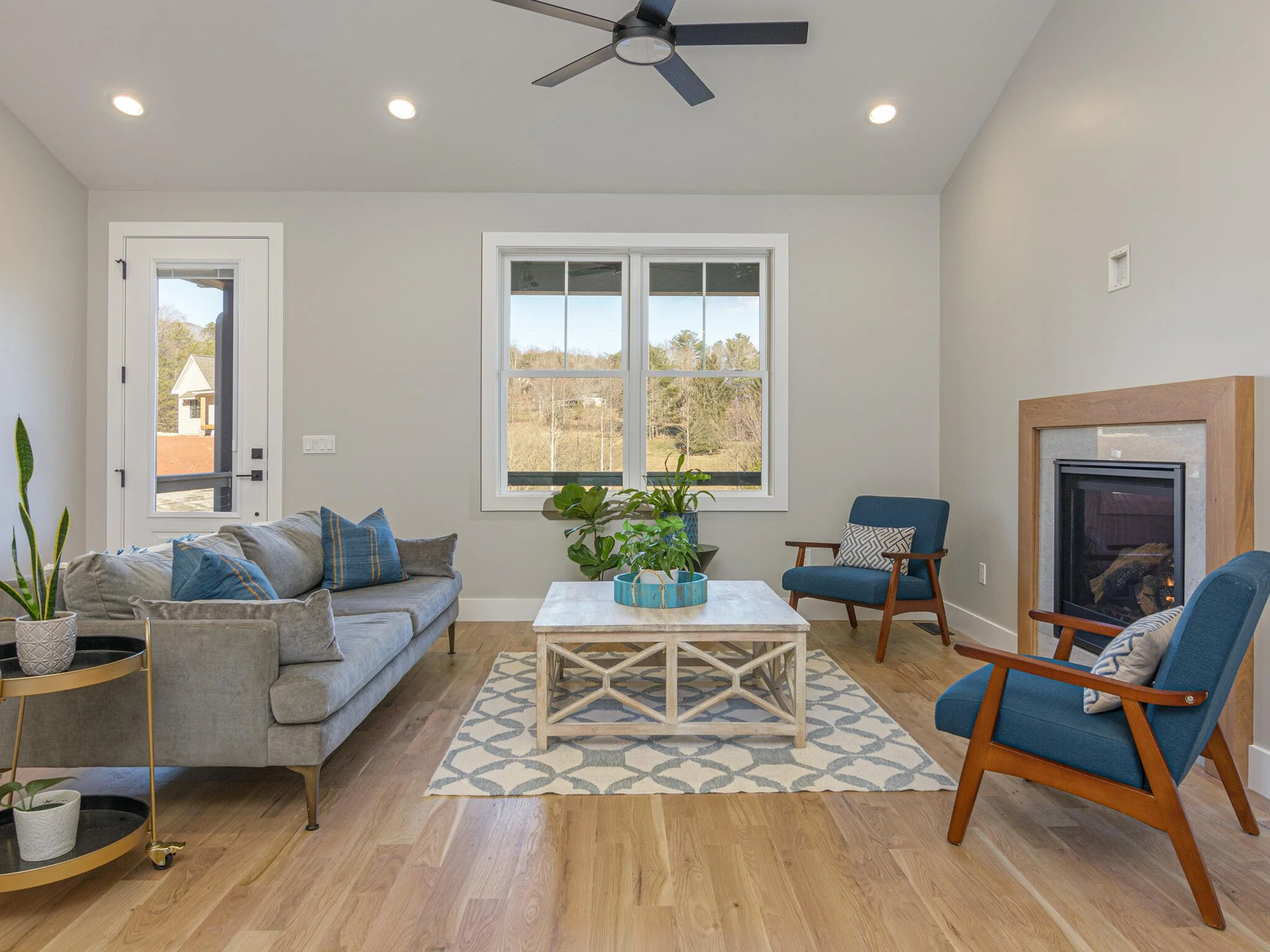 Living room with gray sofa, blue armchairs, a white coffee table, potted plants, a fireplace, and large windows showing a scenic outdoor view.