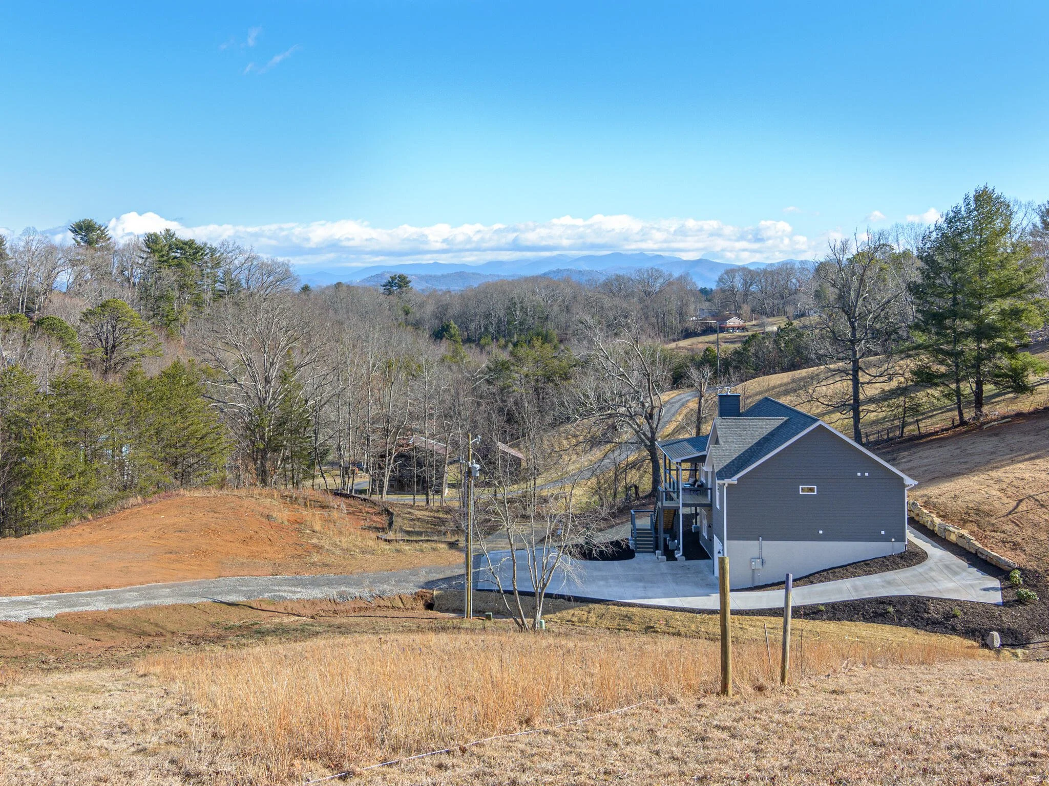 A newly built gray house with a black roof, surrounded by a concrete driveway and a landscaped yard, on a hilly, rural landscape with bare trees, distant mountains, and a partly cloudy blue sky.