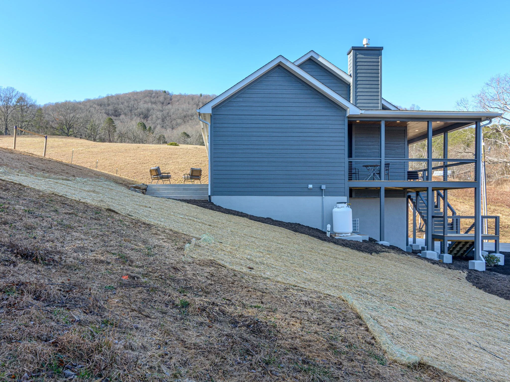 The image shows a modern two-story house on a hillside with a covered porch and external staircase, with a backdrop of trees and hills under a clear blue sky.