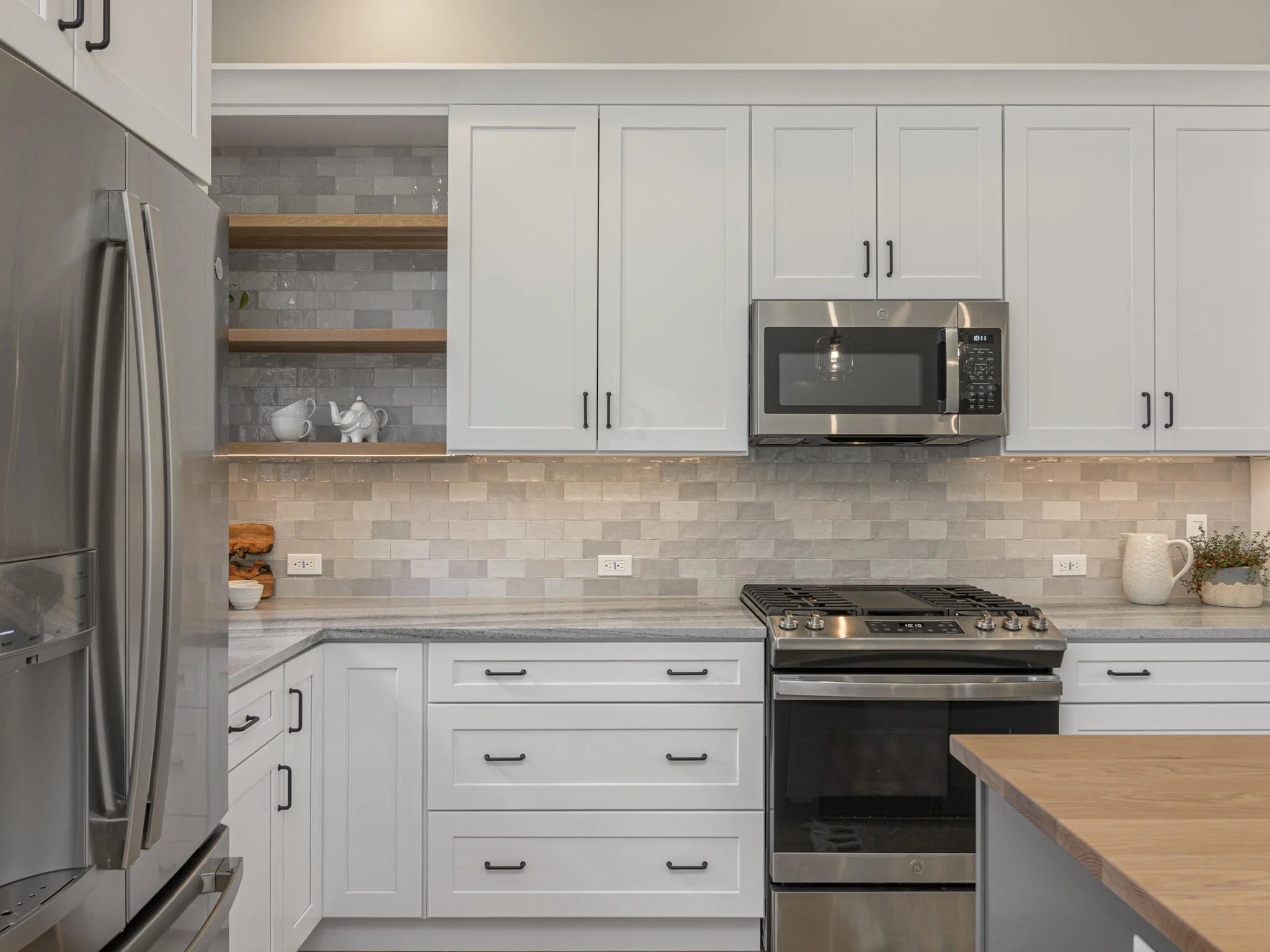 A modern kitchen with white cabinets, stainless steel appliances, open wooden shelves, a granite countertop, gray tile backsplash, and decorative items including tea sets, a wooden tray, and a potted plant.