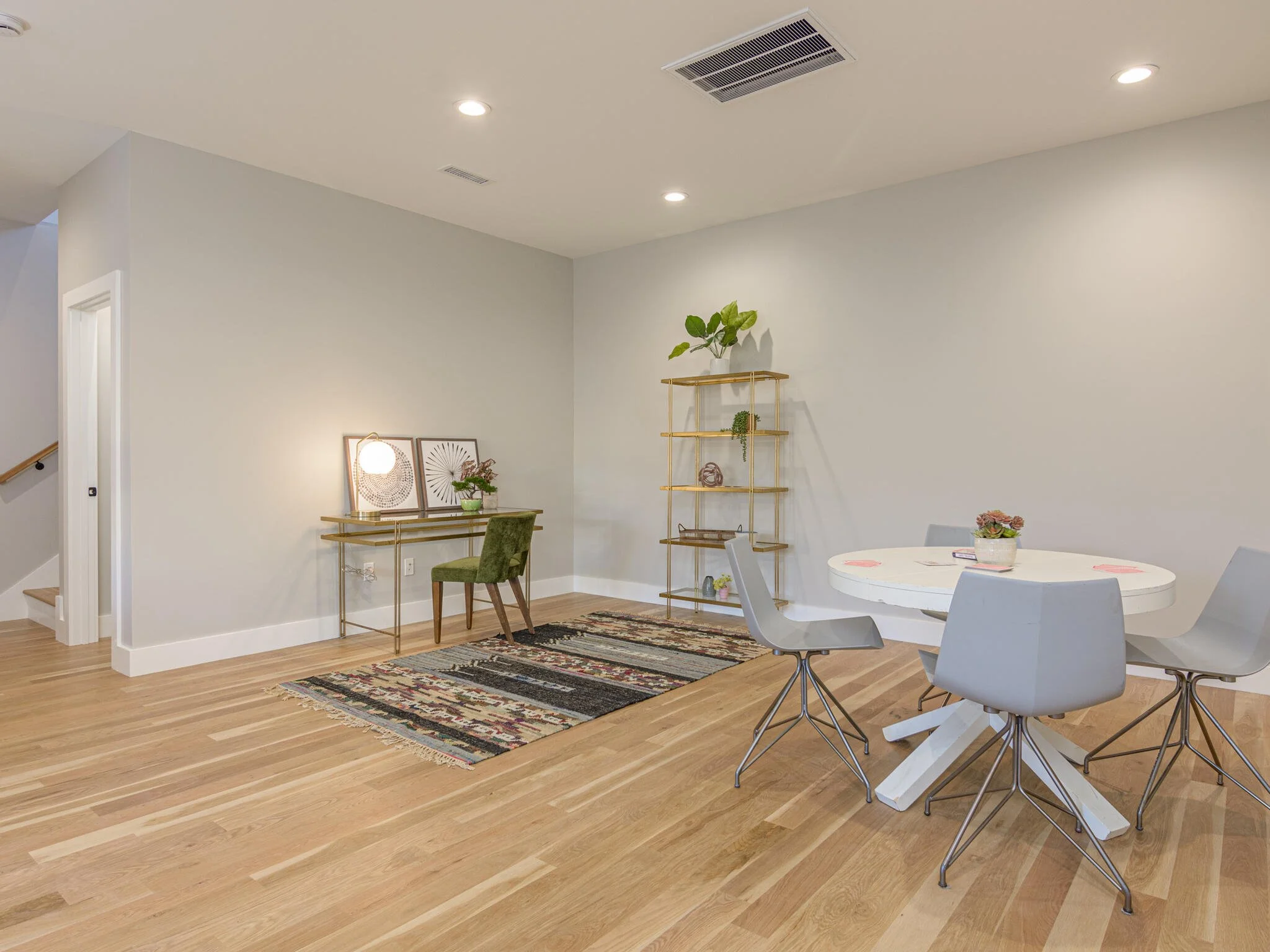 Open living space with a round white table surrounded by four gray chairs, a colorful rug on the wooden floor, a small desk with a green chair, a shelf with plants, and a decorative side table with artwork and a lamp.