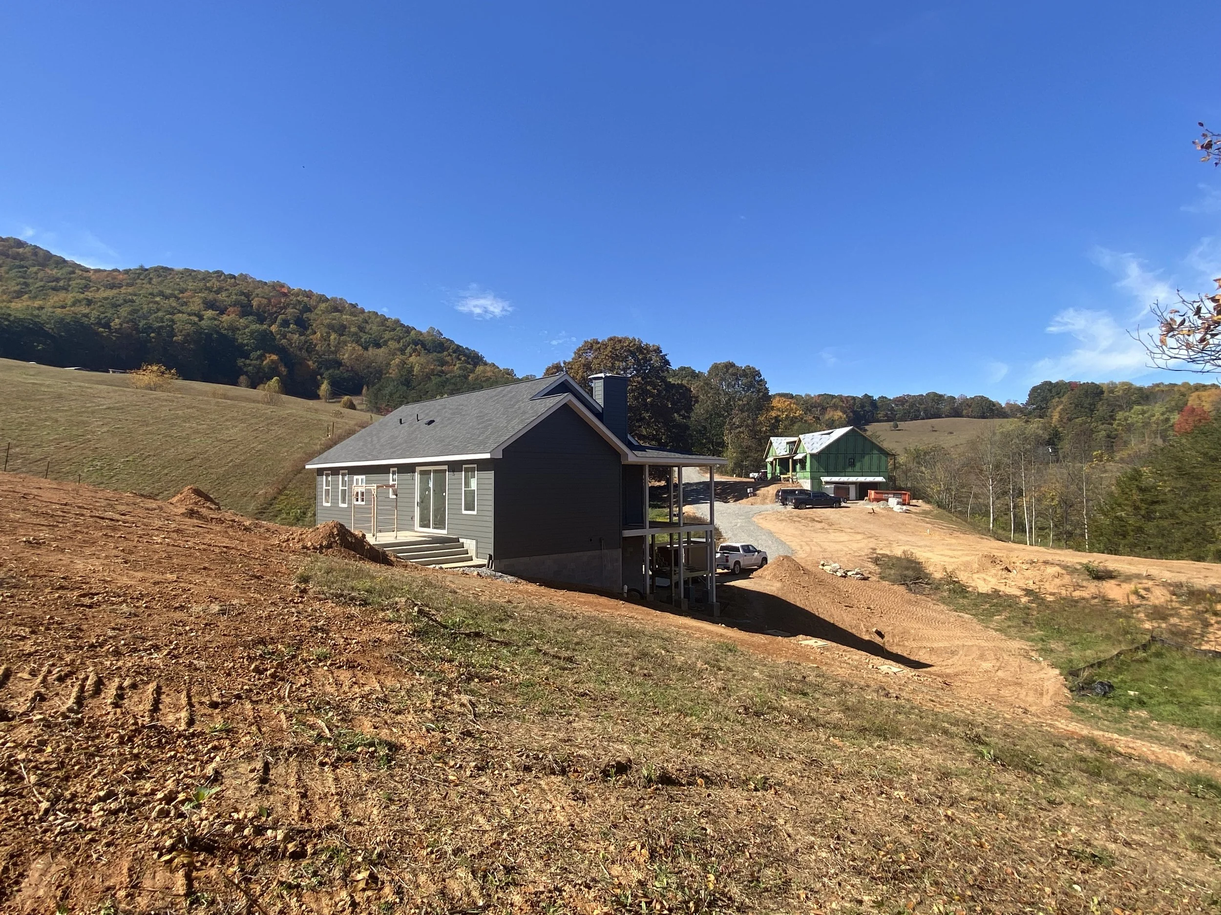 Construction site on a hillside with two houses under construction, surrounded by cleared land and a distant forested hill under a bright blue sky.