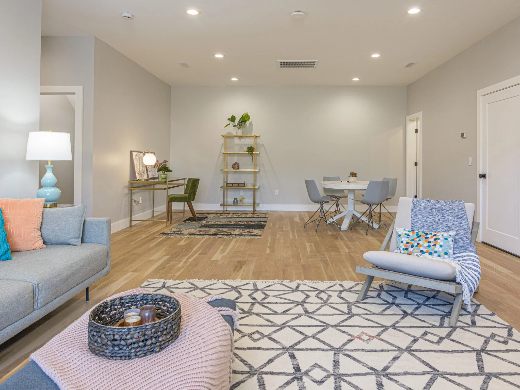 Living room with light-colored walls, hardwood flooring, and modern furniture, including a gray sofa with colorful pillows, a white chair with a throw blanket, a round dining table with gray chairs, a gold shelving unit, a small desk with a green cha