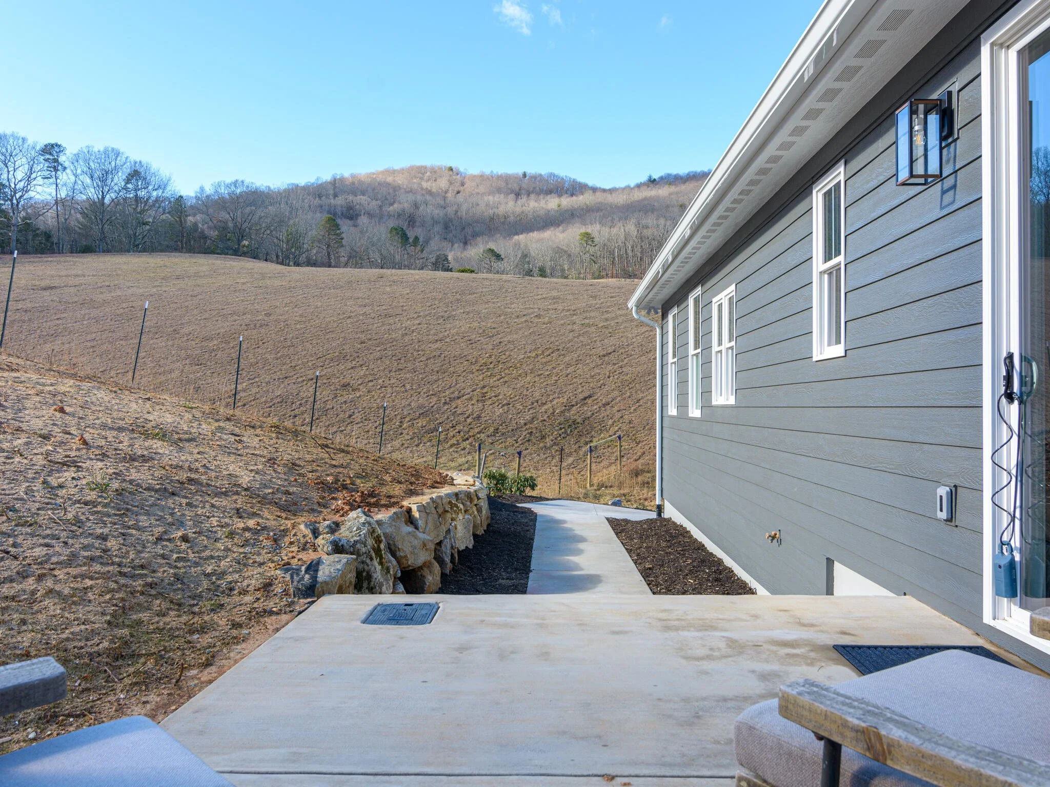 Backyard with a concrete patio next to a gray house, with a view of a hillside and trees in the distance.