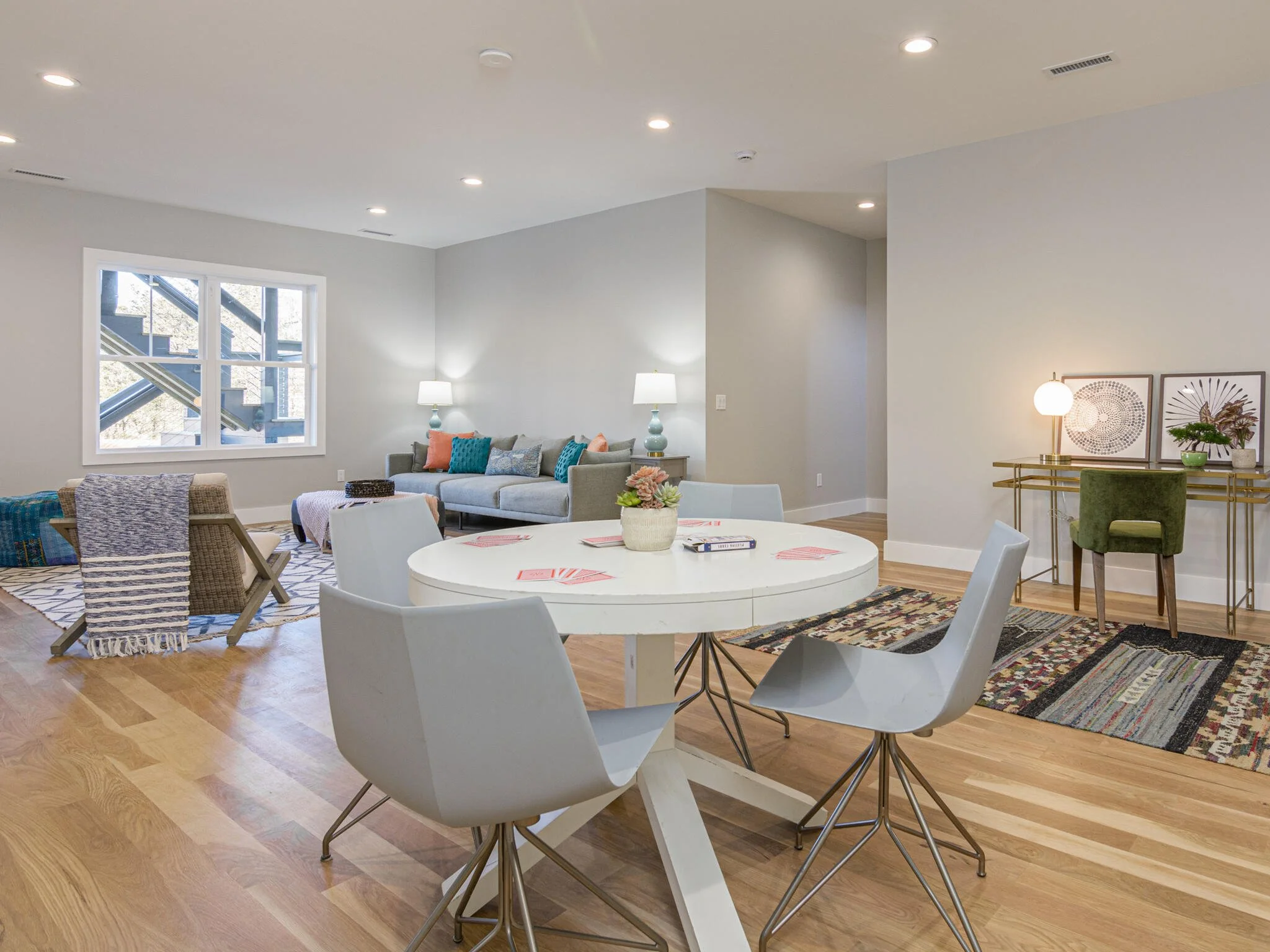 Open-concept living and dining area with hardwood floors, gray walls, and modern furniture. A round white dining table with four white chairs is in the foreground, decorated with a small plant and red placemats. In the background, there is a gray sof