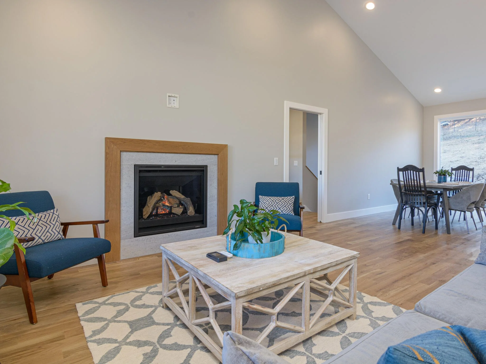 Living room with a fireplace, blue chairs, a wooden coffee table with a plant, a patterned rug, and a dining area with a round table and chairs near a large window showing an outdoor hill.