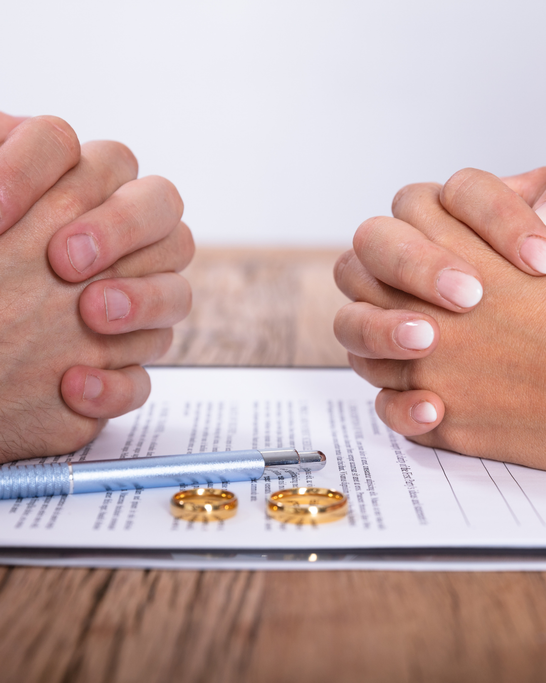 Close-up of two people with clasped hands over a document containing text, with two gold wedding rings, a blue pen, and a wooden surface.