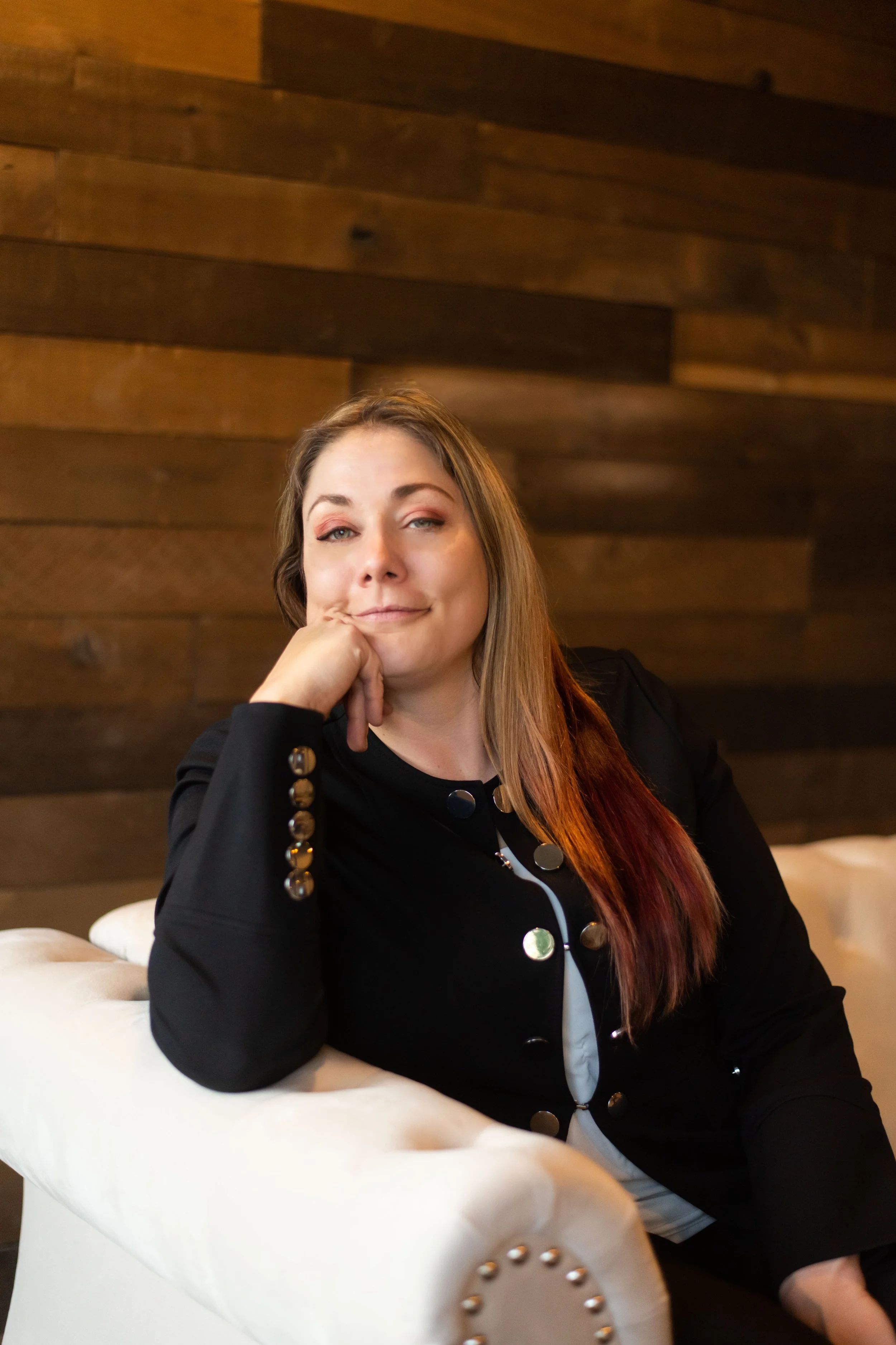 A woman with long hair sitting on a white couch with a wood-paneled wall in the background, resting her chin on her hand and looking at the camera.