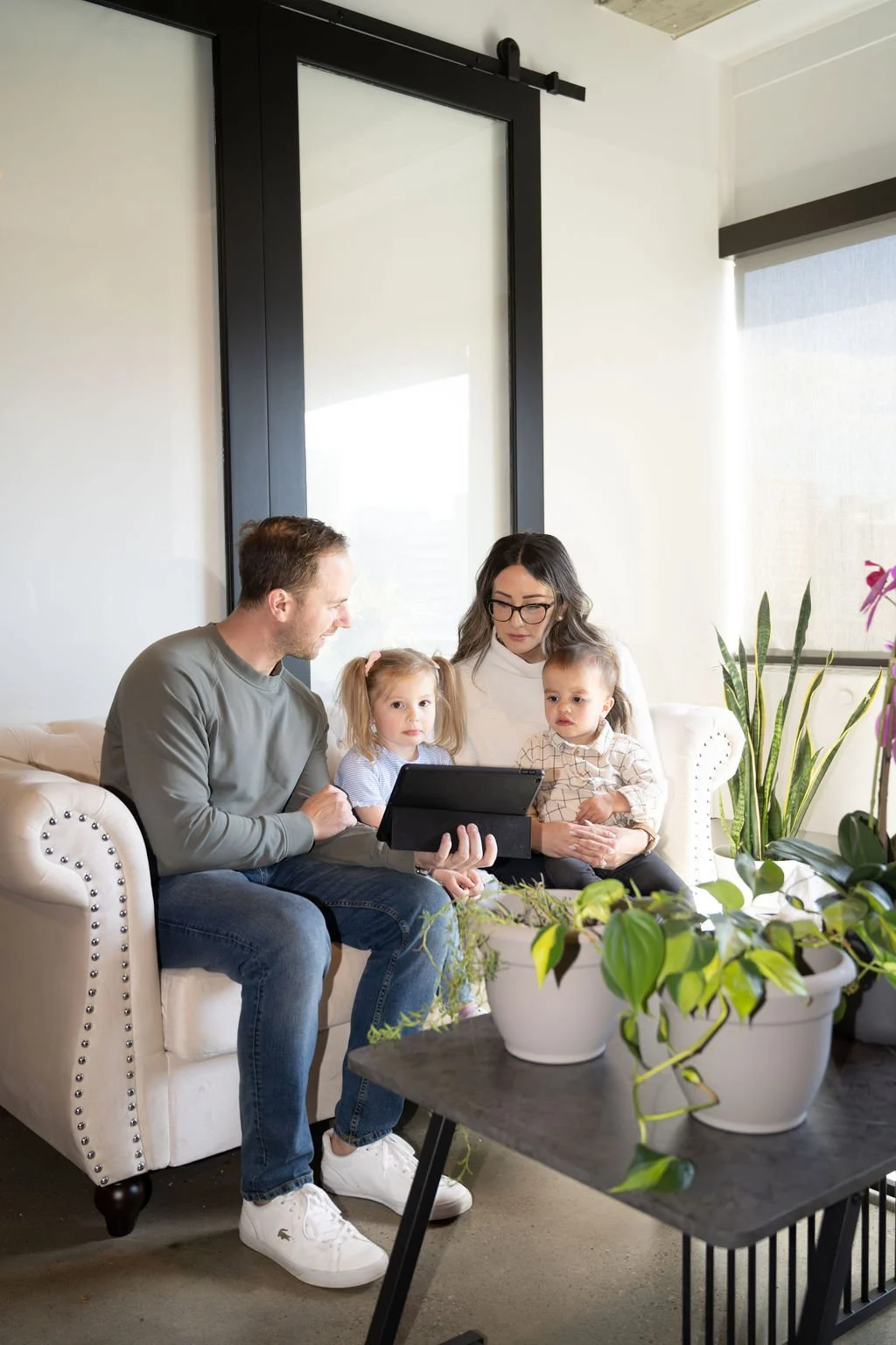 A family of four sitting on a white couch, looking at a tablet, in a room with large windows and indoor plants.