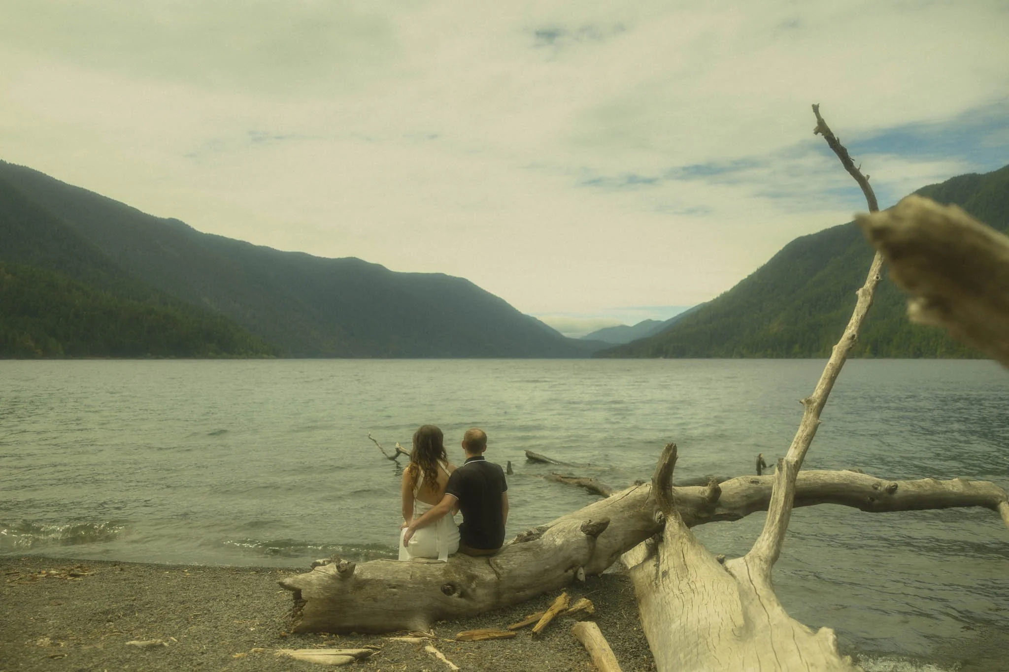 a couple sitting on a piece of driftwood on the shore of Lake Crescent