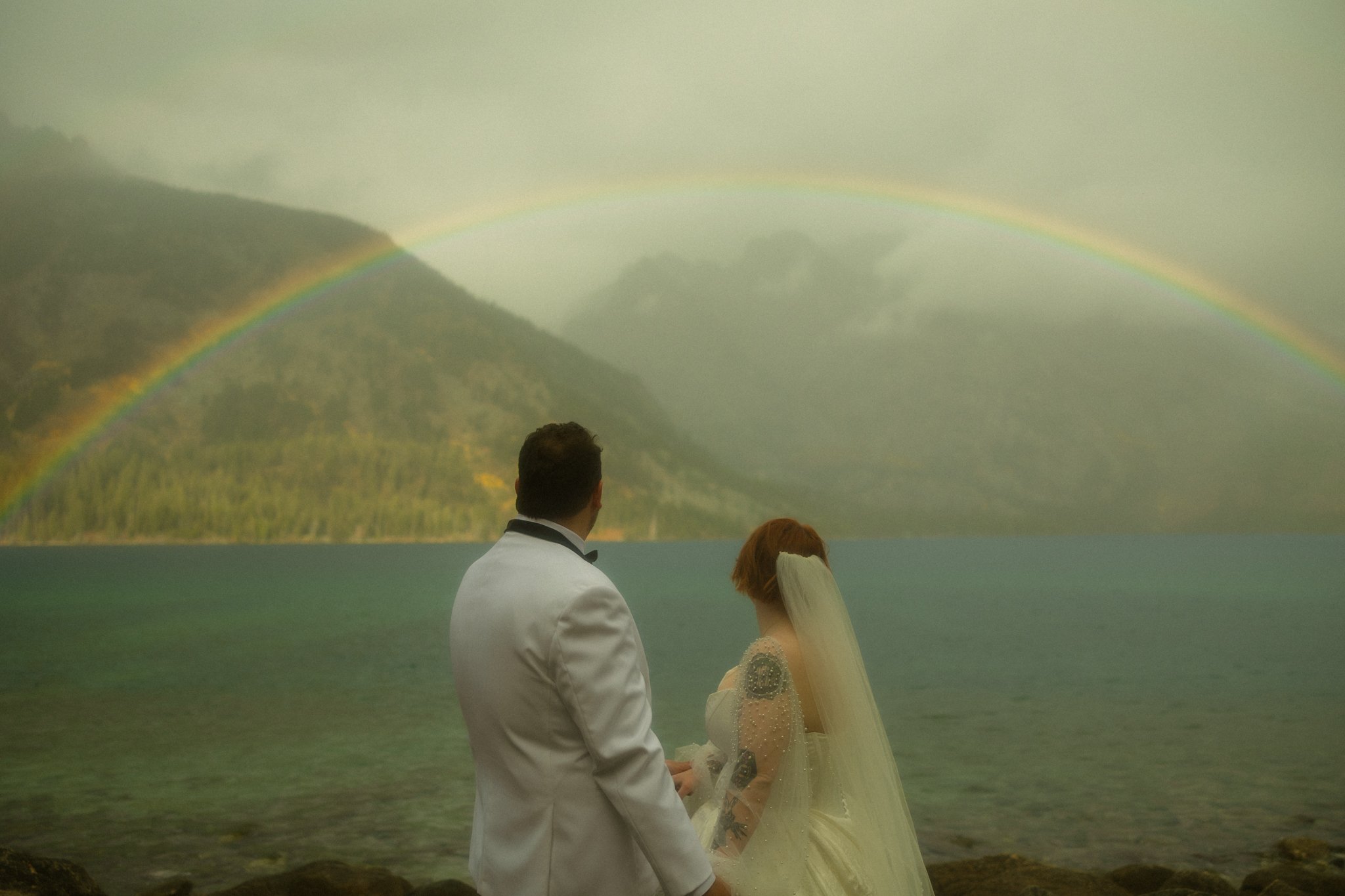 an elopement couple standing on a lakeshore with a rainbow in the background