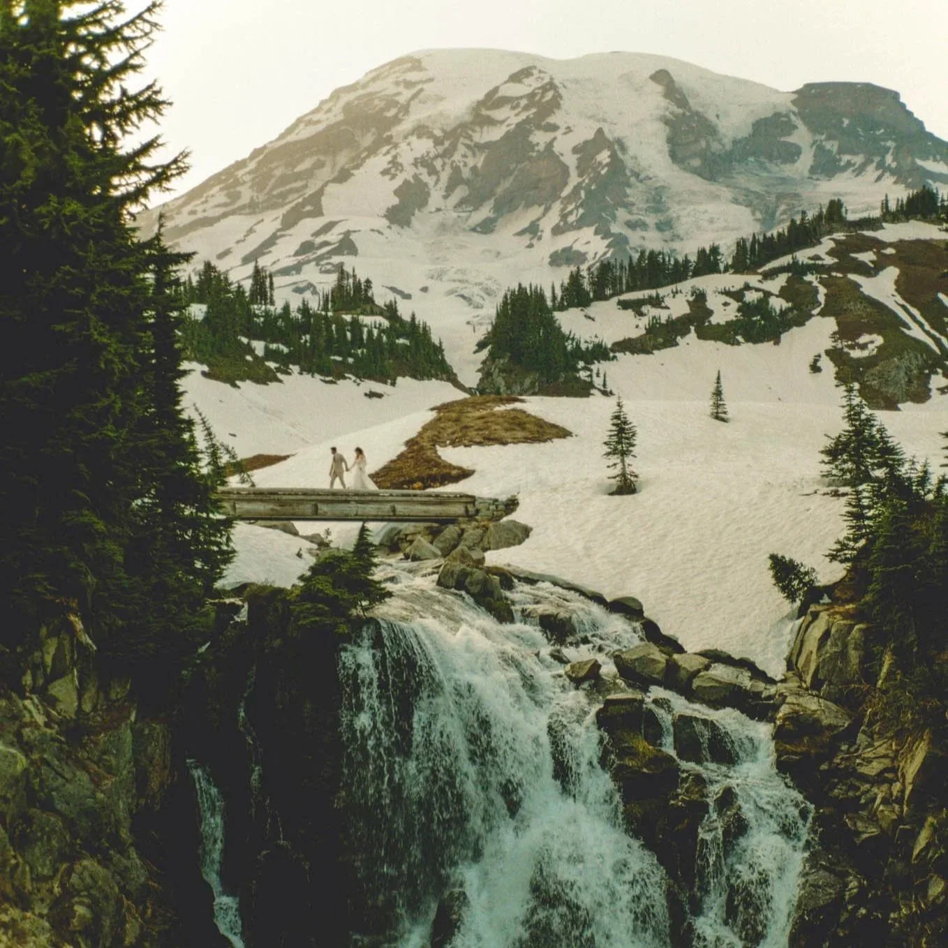 an elopement couple walking across a waterfall bridge in snowy Mt Rainier