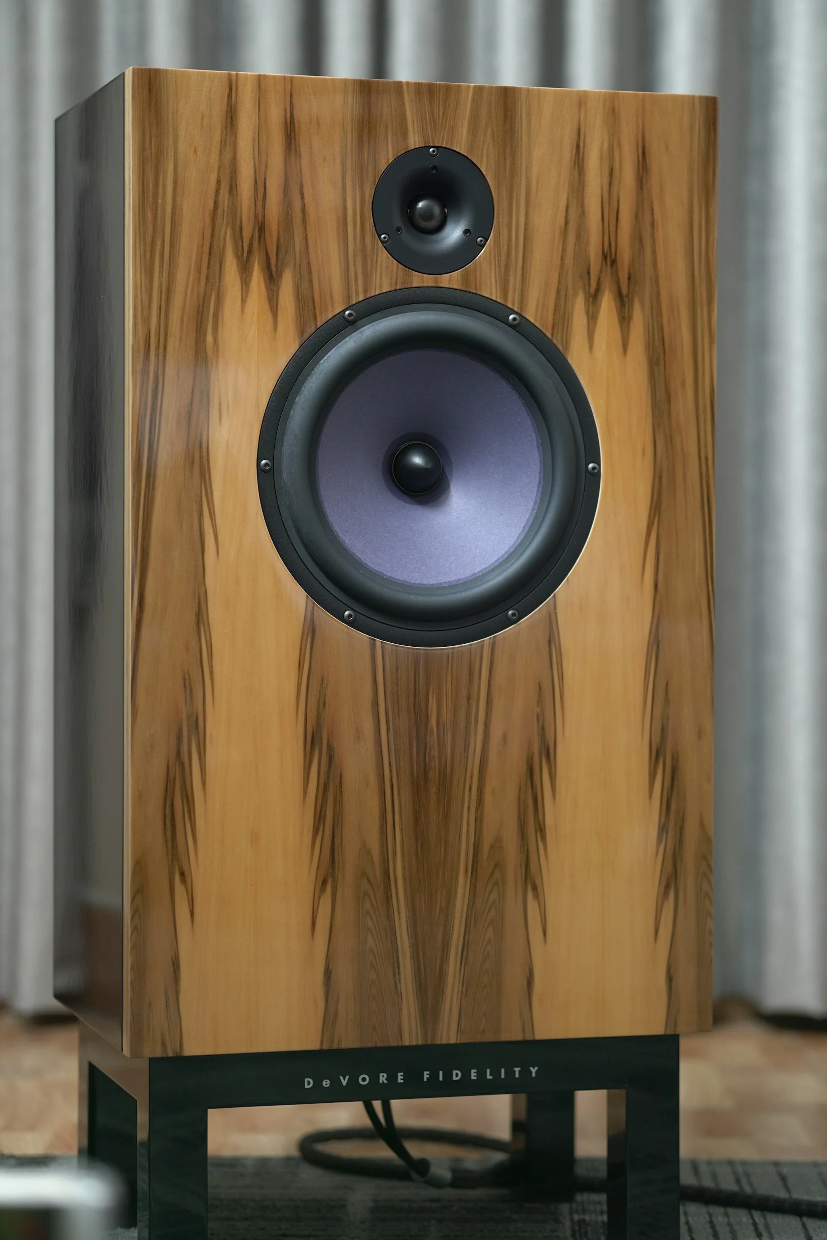 A large wooden bookshelf speaker with a black base. The speaker has a wood grain finish and features two black drivers, a smaller one above and a larger one below. The brand name 'DeVORE FIDELITY' is visible on the black base of the speaker.