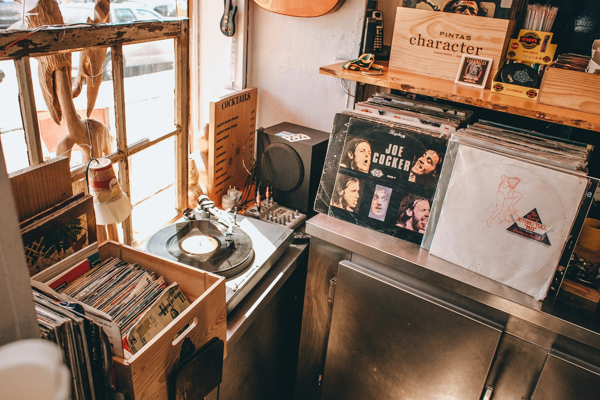 A vintage record shop with vinyl records, a turntable, and a small stereo system. Wooden shelves hold more records and a sign reading 'character.' The shop has a window with sunlight streaming in, and various posters and decorations adorn the walls.