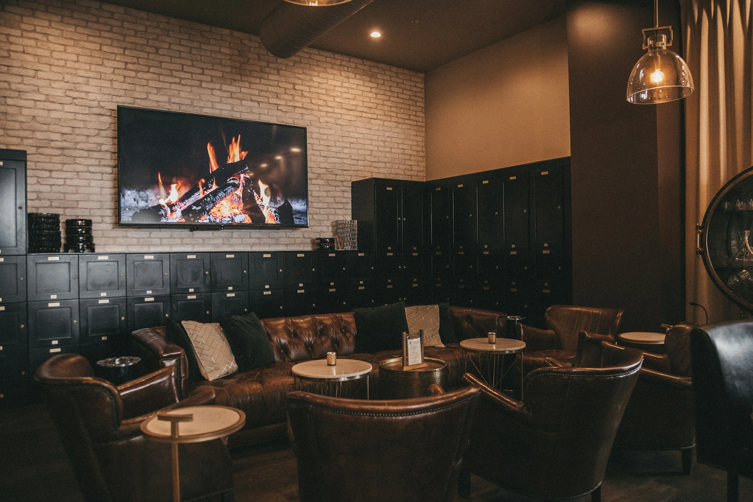 Cozy lounge with leather armchairs and a long leather sofa, small round tables, black lockers lining the wall, a large wall-mounted TV showing a fireplace, brick wall background, and warm lighting.