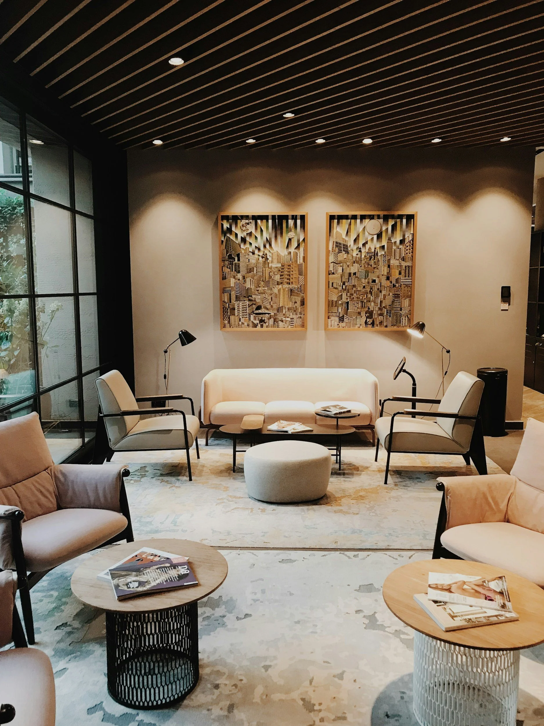 A cozy hotel lobby with modern decor, including white and beige upholstered chairs, a white sofa, a round ottoman, and wooden side tables with magazines. The wall behind features two framed abstract cityscape art pieces illuminated by spotlights, and a large window on the left side offers natural light.